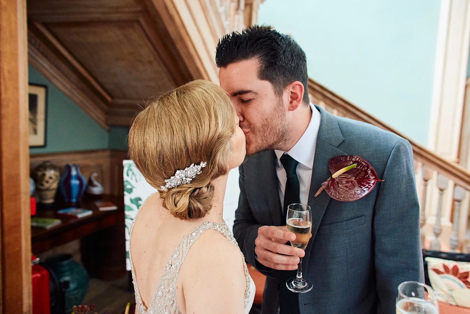 A newly married couple share an intimate kiss indoors, with the groom in a grey suit holding a glass of champagne while the bride wears an embellished gown and a crystal hair comb.