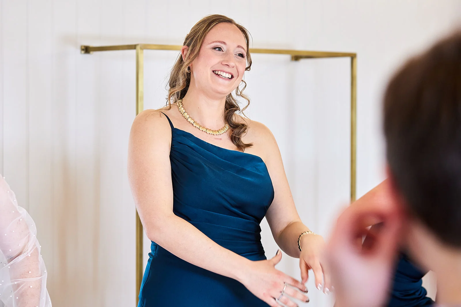 A smiling bridesmaid in a teal satin one-shoulder dress laughs during morning wedding preparations.