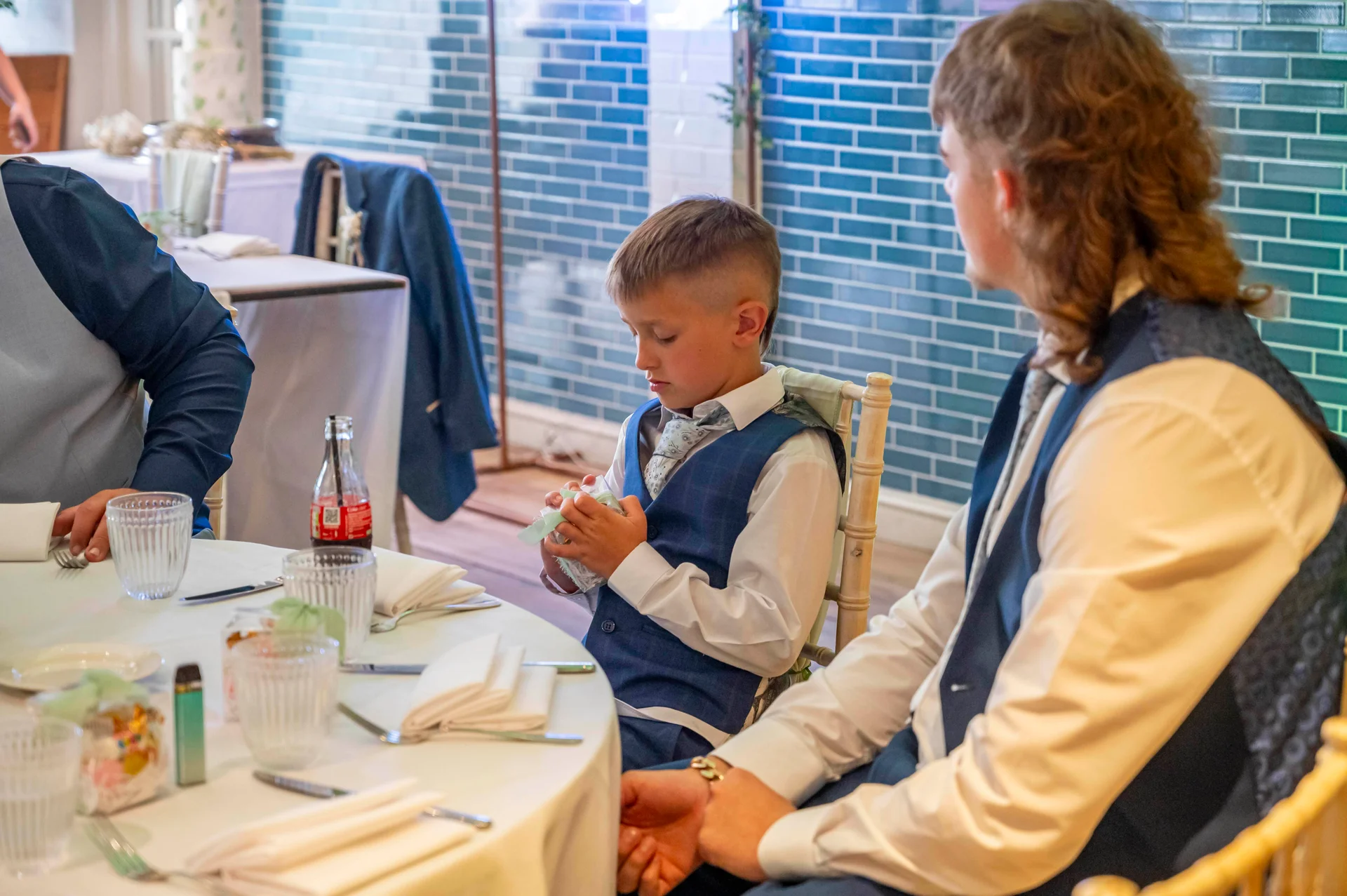 A young page boy and a groomsman sit at a wedding breakfast table indoors with a blue tiled backdrop.
