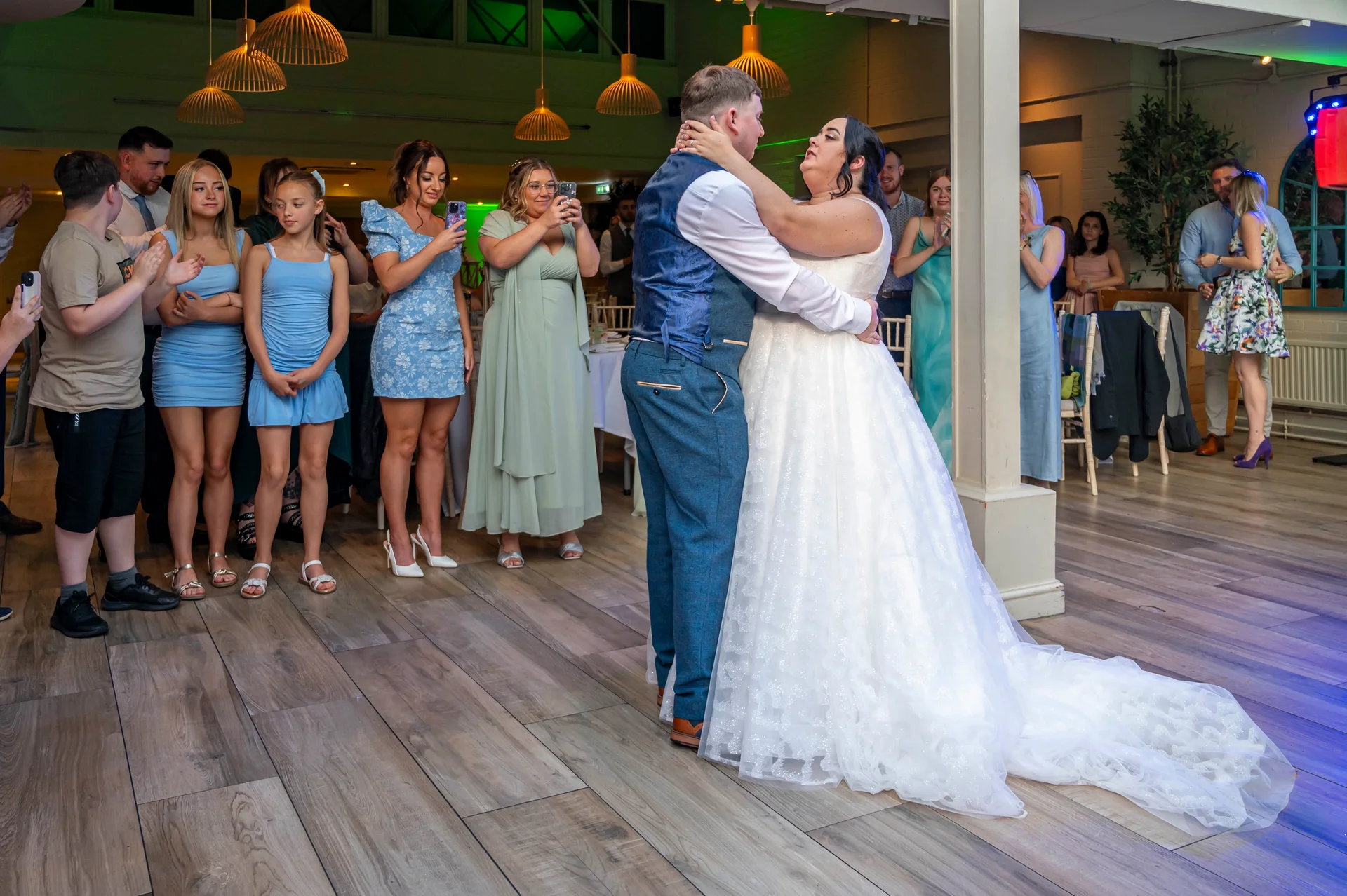 Newlyweds share their romantic first dance on a wooden floor in a warmly lit venue surrounded by watching family and guests.