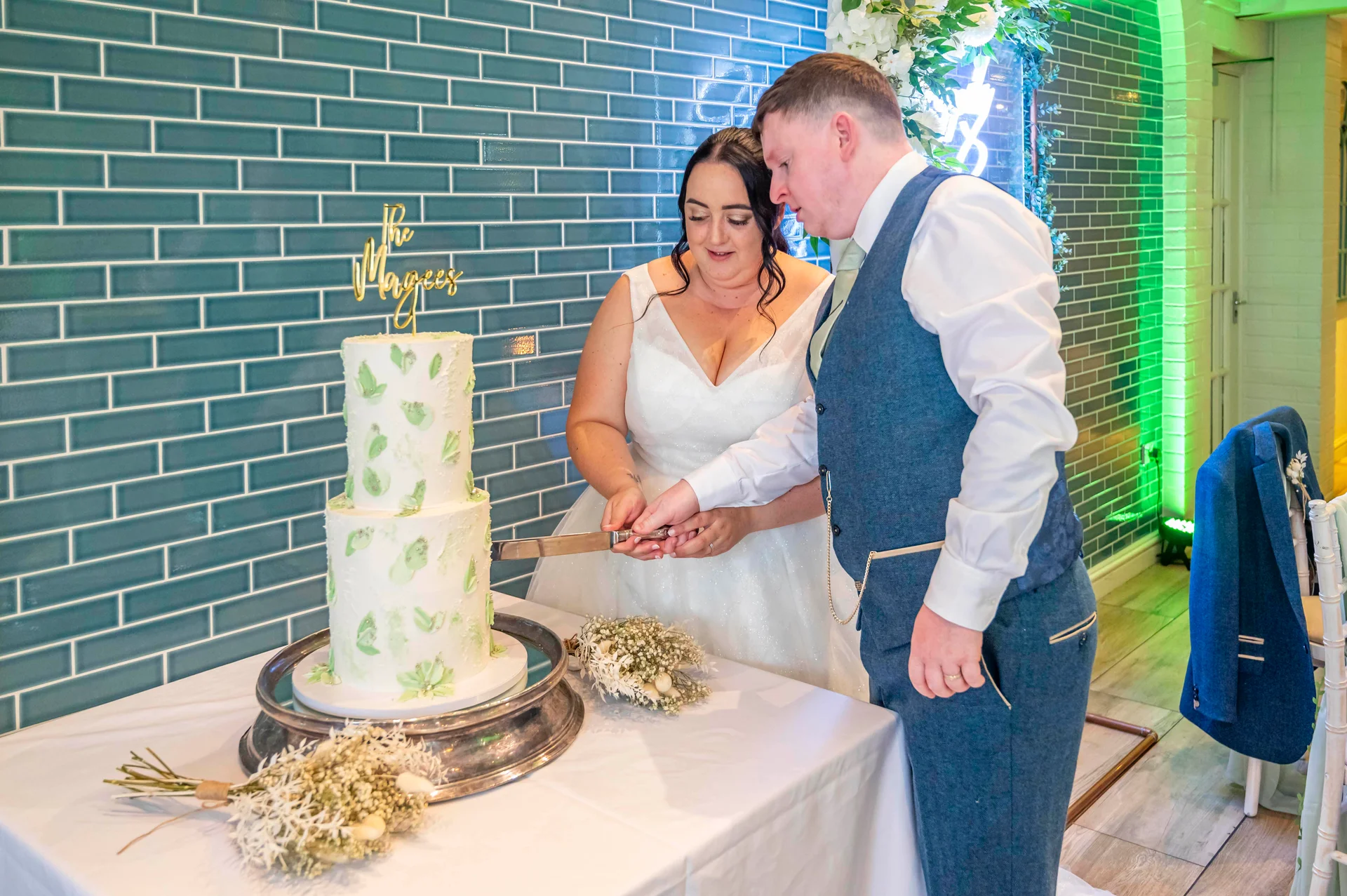 The bride and groom cut into a two-tier green and white buttercream wedding cake against an industrial blue tiled wall.