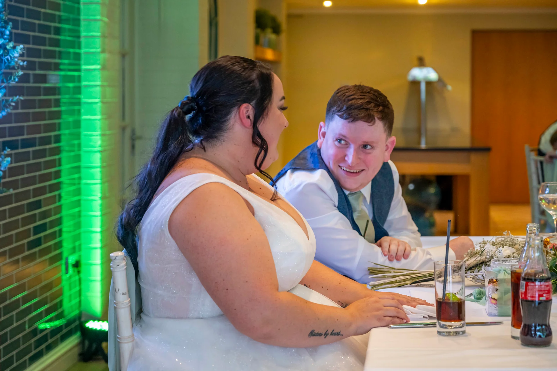 A bride with a dark ponytail and a groom in a blue waistcoat share a joyful, laughing glance while seated at the top table during their wedding breakfast.