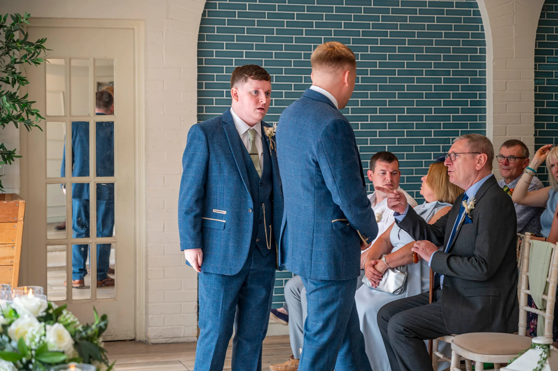Groomsmen in matching blue tweed suits converse near a blue tiled wall while guests look on during a wedding celebration.