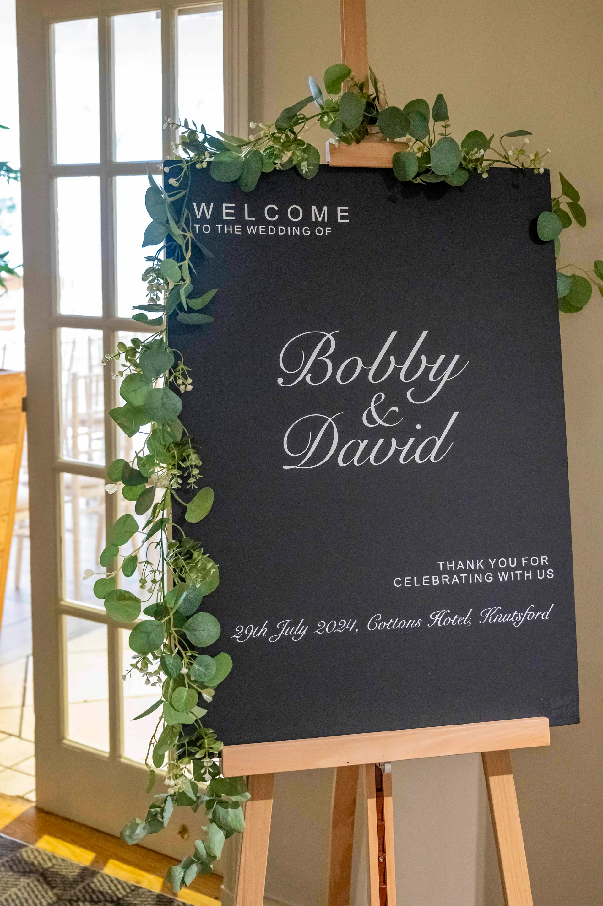 A modern black wedding welcome sign on a wooden easel is elegantly decorated with a lush eucalyptus greenery garland.