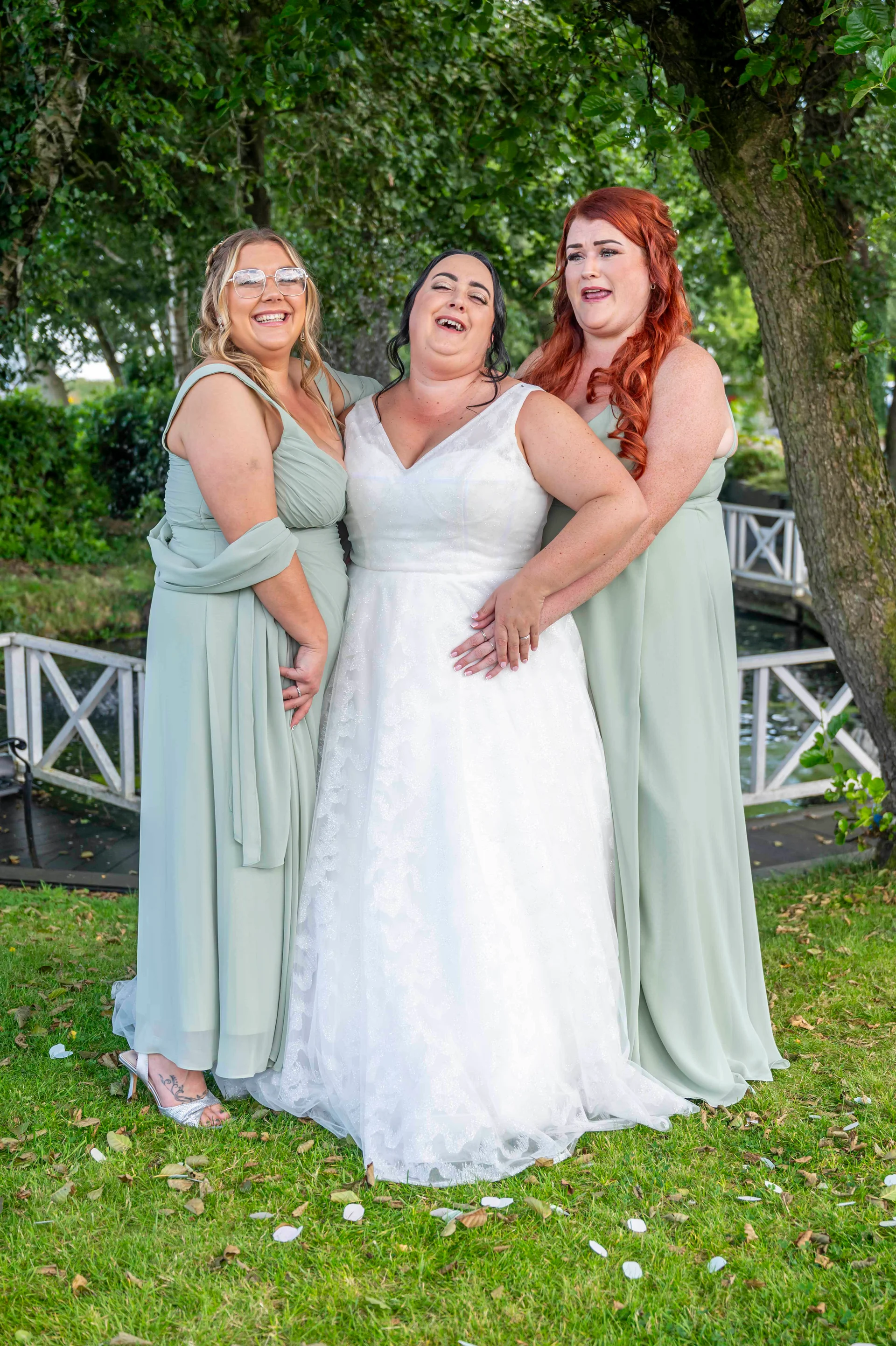 A laughing bride stands between her two bridesmaids wearing sage green dresses outdoors by a white wooden fence and lush trees.