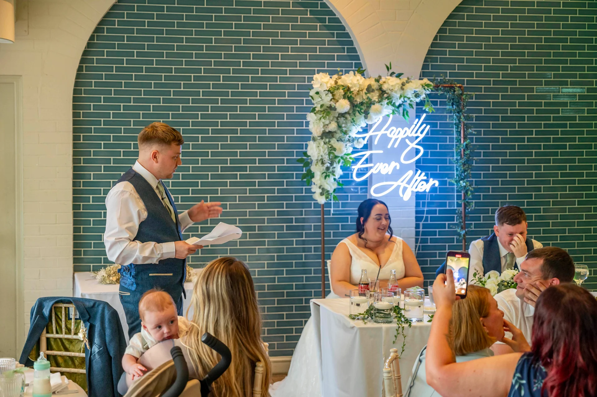 A groom delivers his speech at the top table during a wedding reception, with guests watching and recording on phones in front of a blue tiled wall and illuminated neon sign.