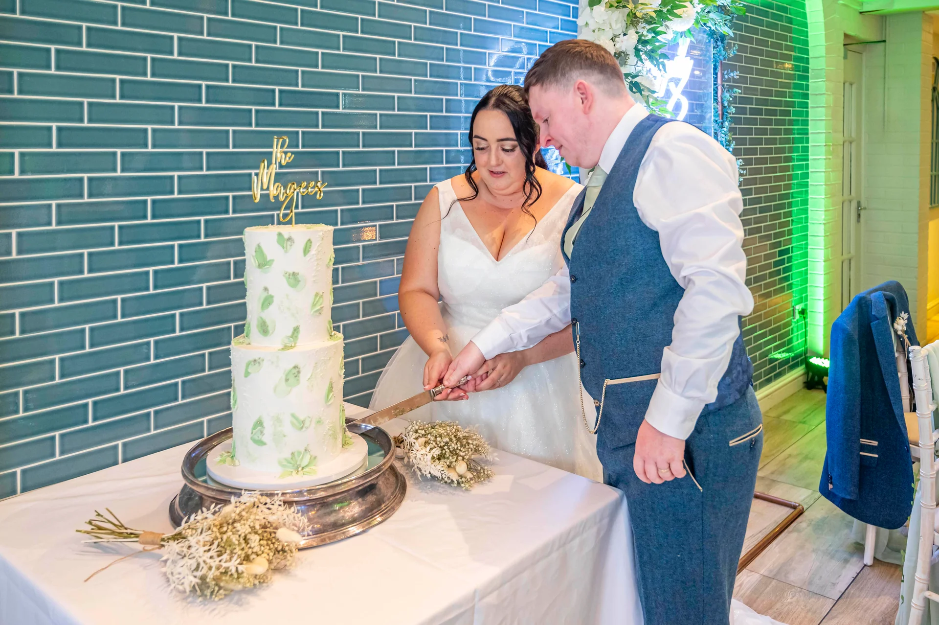 The bride and groom stand together to cut a two-tier white buttercream wedding cake decorated with green leaf details against a teal tiled wall.