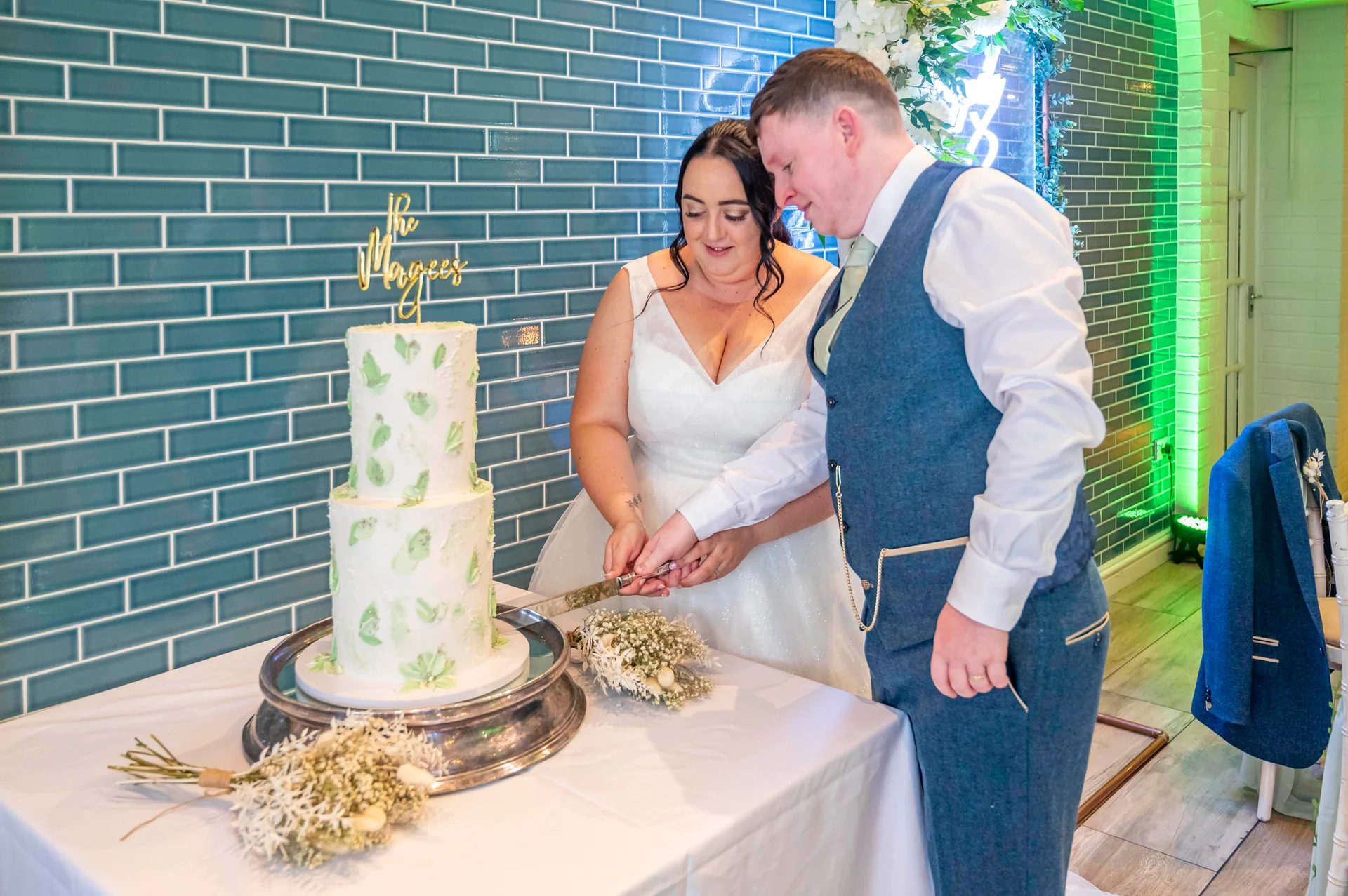A newlywed couple smiling as they cut a two-tier white wedding cake decorated with green leaf accents in front of a teal tiled wall.