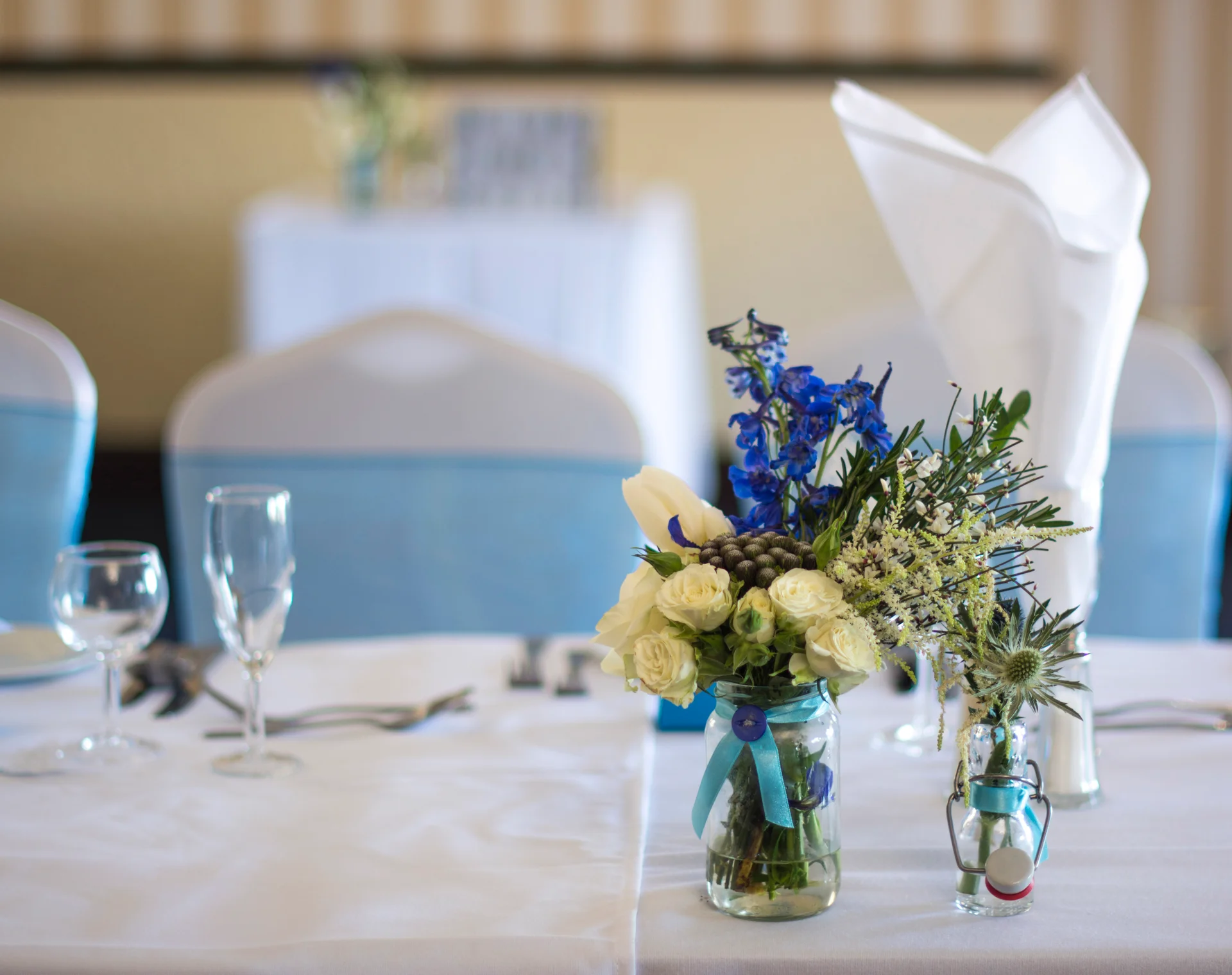 A close-up view of a wedding reception tablescape featuring a mason jar filled with white roses, blue delphiniums, and thistles alongside a folded white napkin.