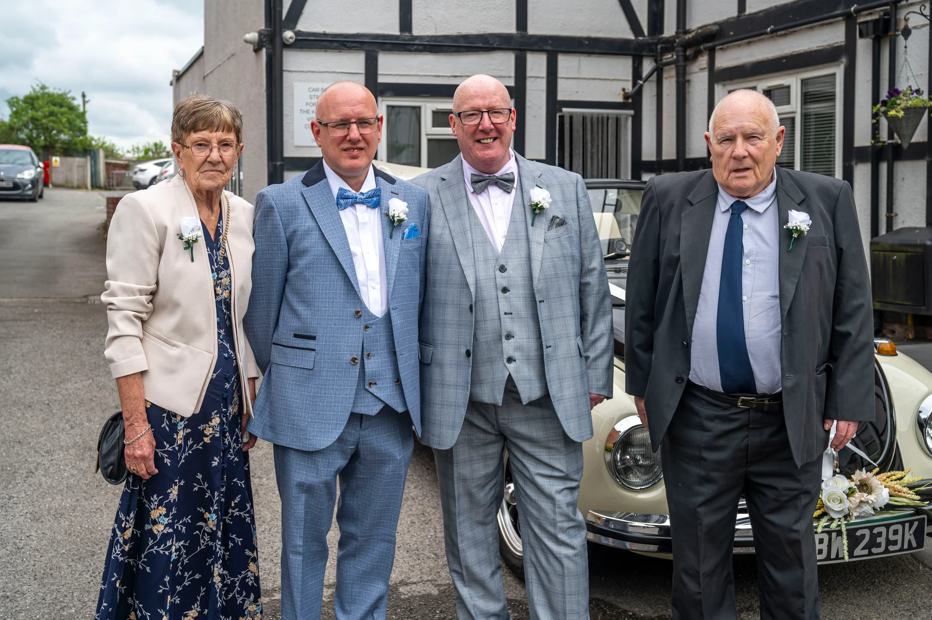 A family group posing outside next to a vintage wedding car, featuring two men in tailored suits with buttonholes, an older woman in a floral dress, and another gentleman in a dark suit and tie.