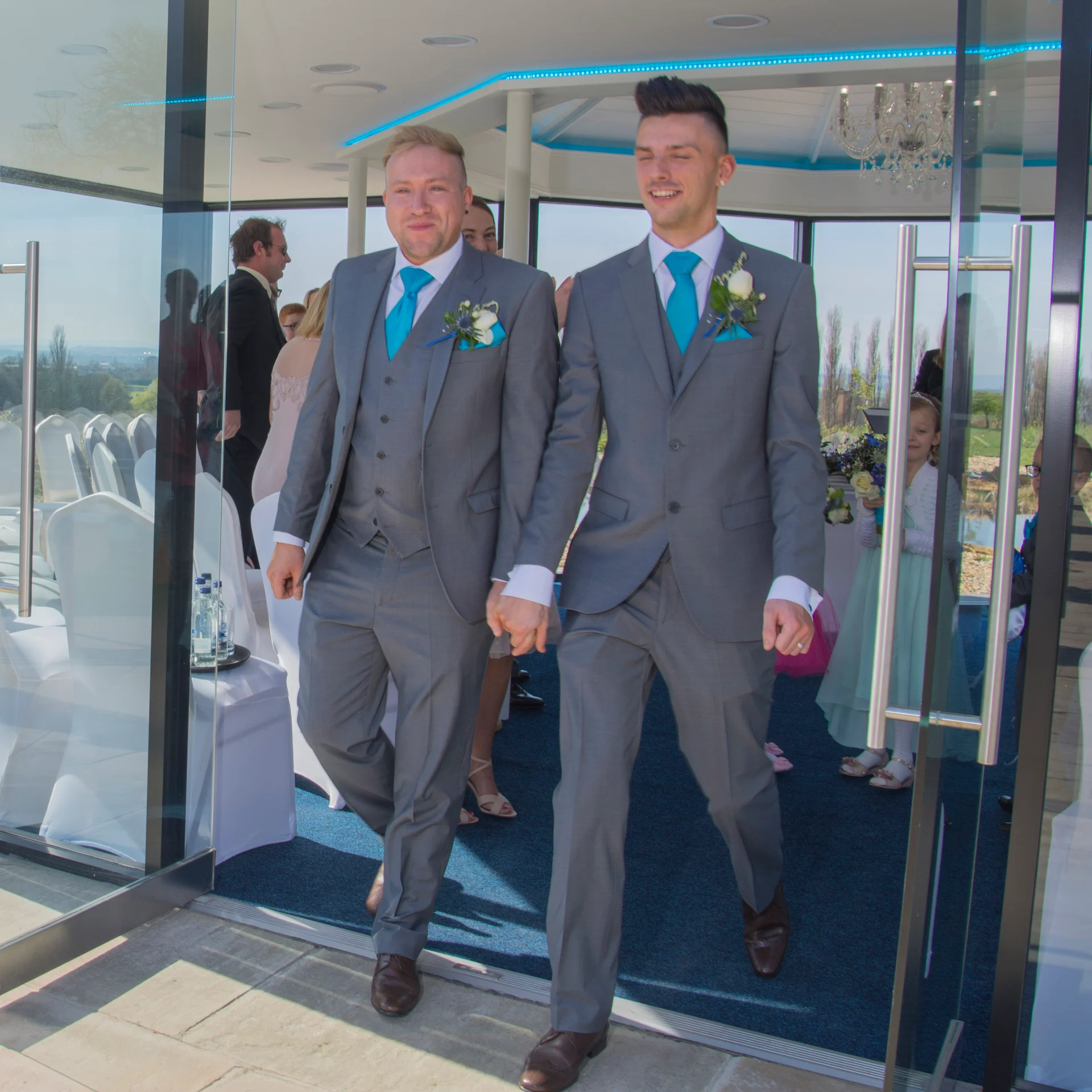 Two smiling grooms in matching grey suits and bright blue ties walk hand in hand out of a glasshouse ceremony venue.