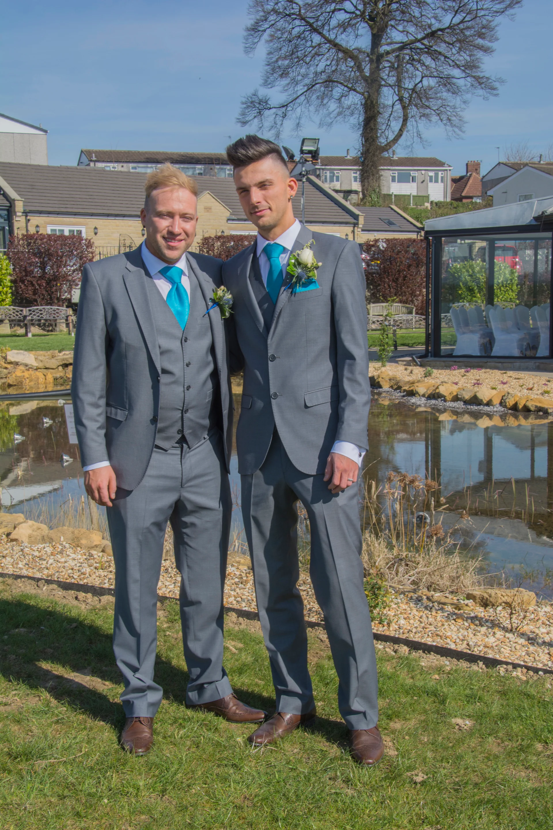 Two grooms stand side by side wearing matching grey suits and bright blue ties by a tranquil outdoor pond on a sunny wedding day.