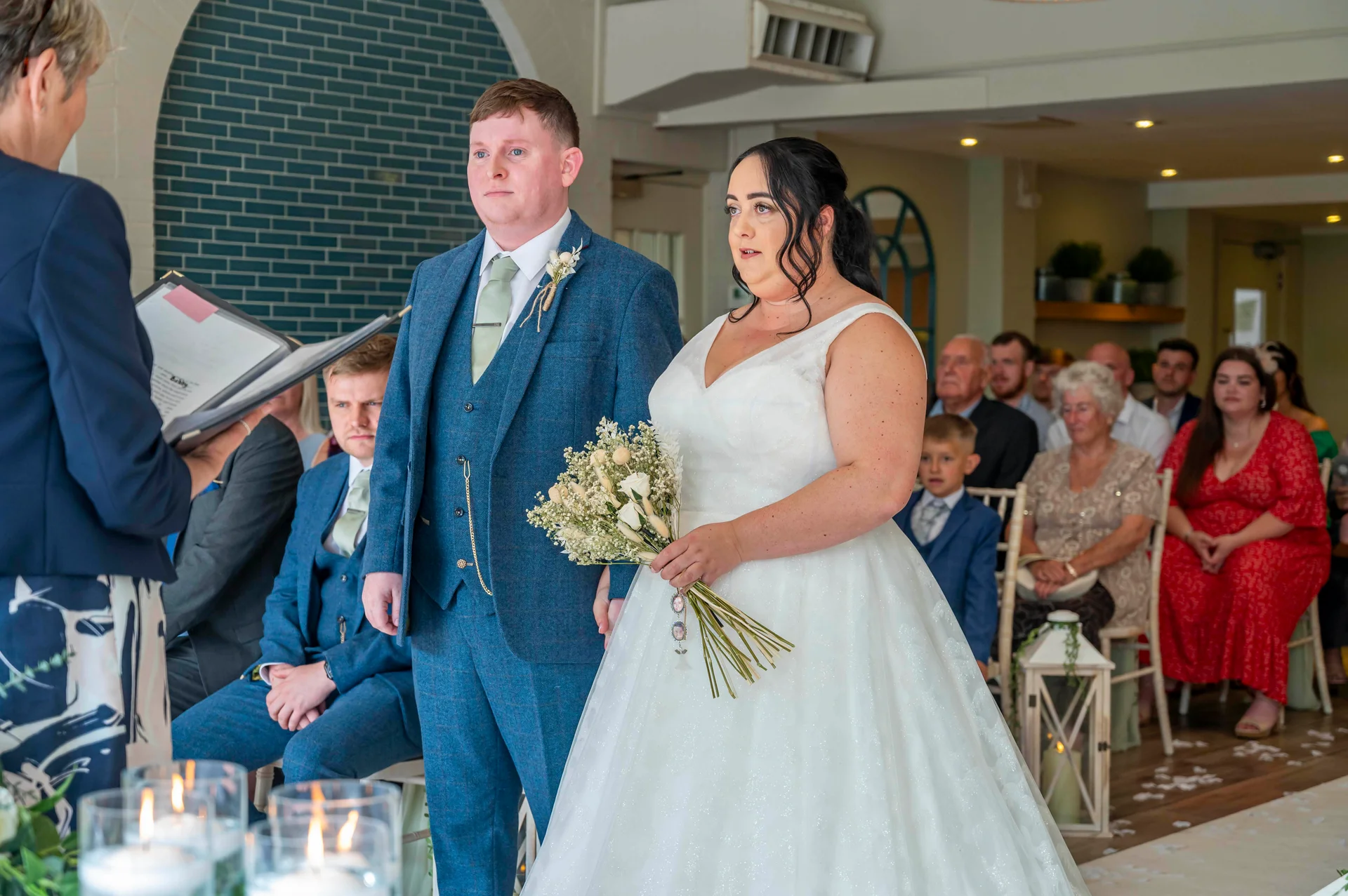 A bride holding a bouquet and groom in a blue tweed suit stand attentively during their civil ceremony as guests watch.