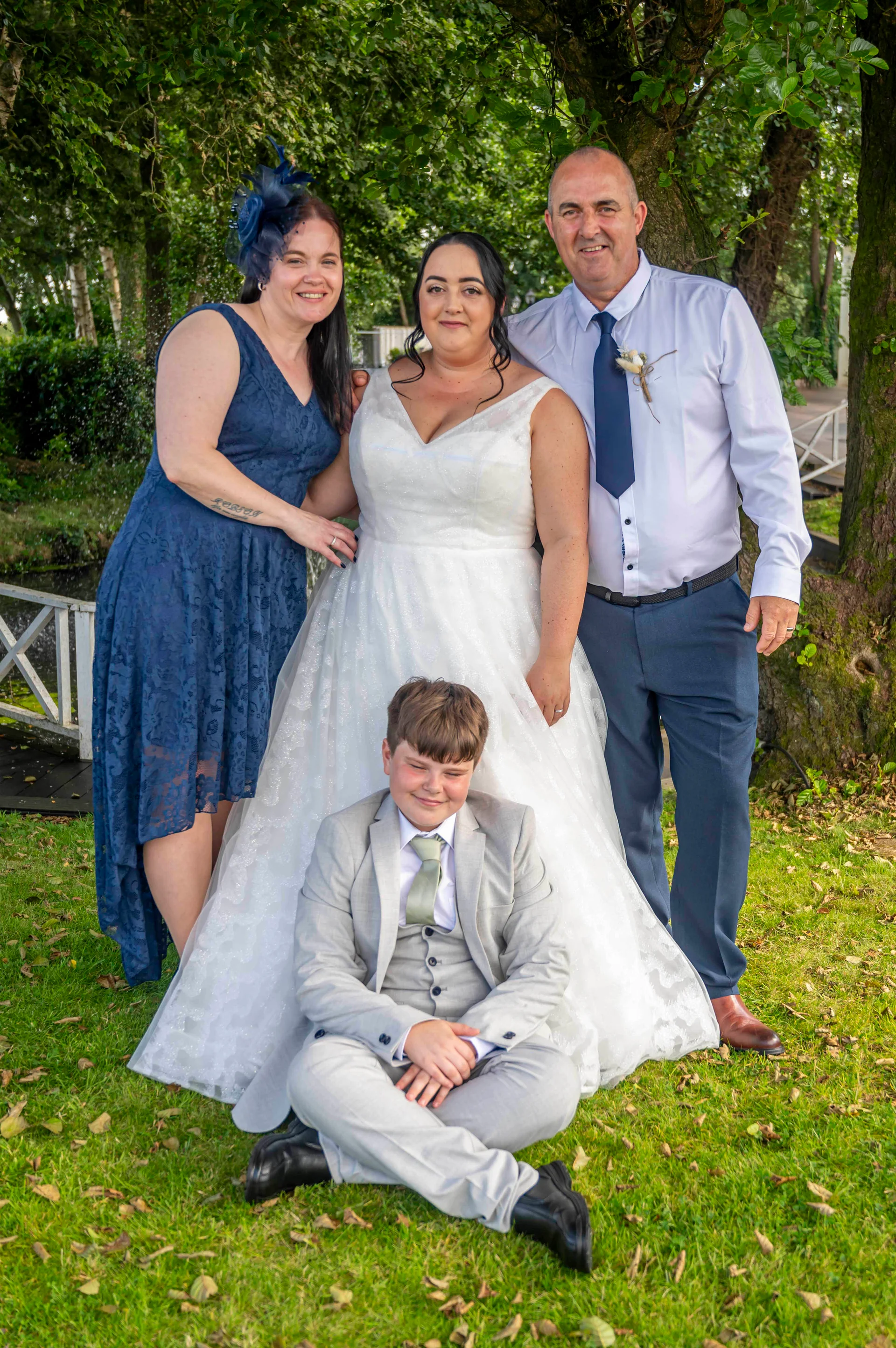 A bride in a white A-line gown poses outdoors on the grass with her family, including a bridesmaid in a blue dress, a man in a light blue shirt with a tie, and a young boy sitting cross-legged in a grey suit.