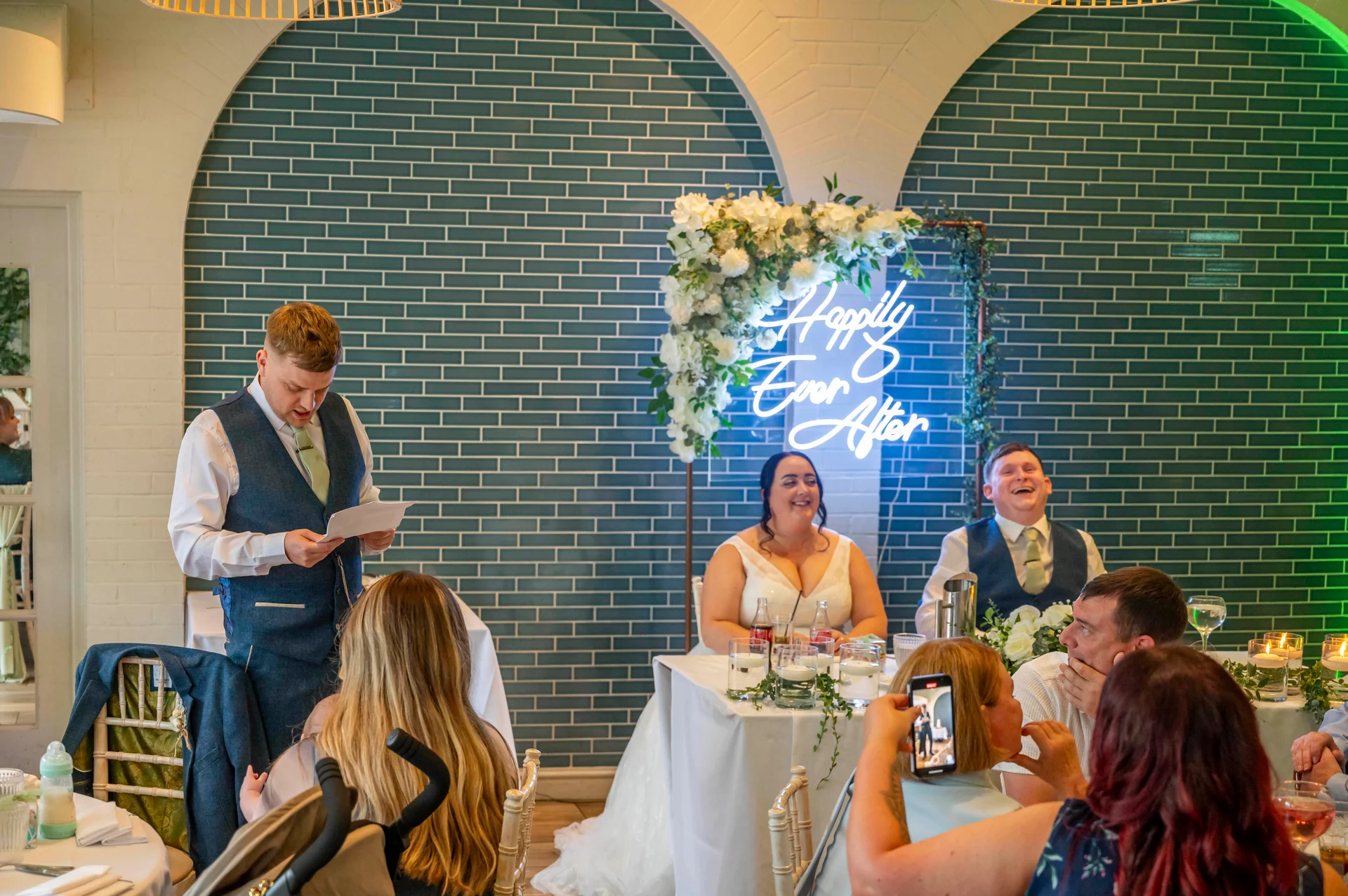 A groomsman delivers a speech at the top table while the bride and groom laugh in front of a blue tiled wall with a neon sign and floral arch.