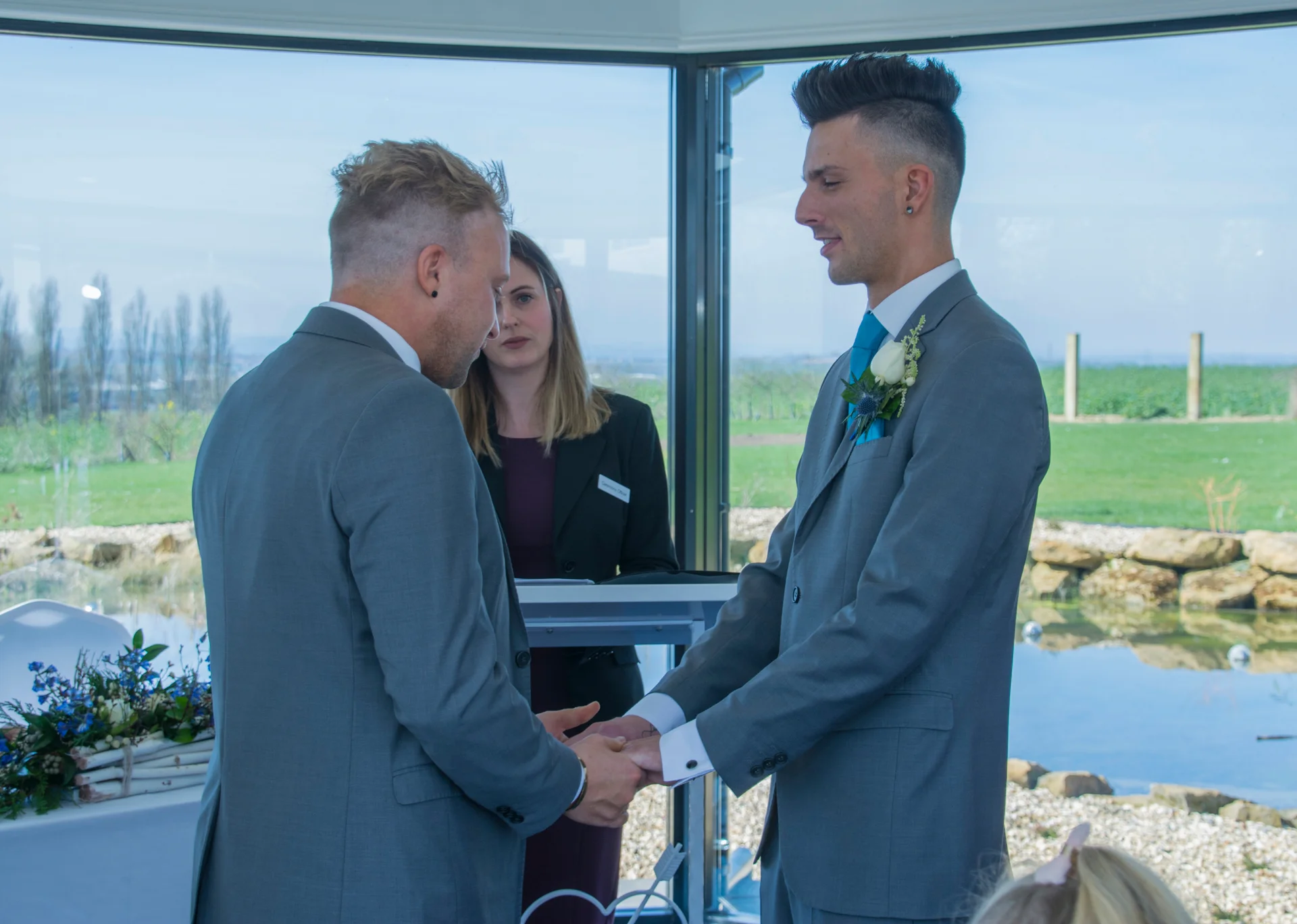 Two grooms in matching grey suits hold hands and exchange vows during a light-filled civil ceremony inside a modern glasshouse venue.