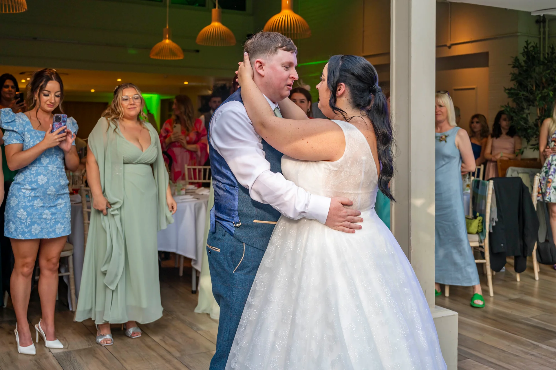Newlyweds share their first dance indoors while wedding guests watch and capture the moment on their phones.
