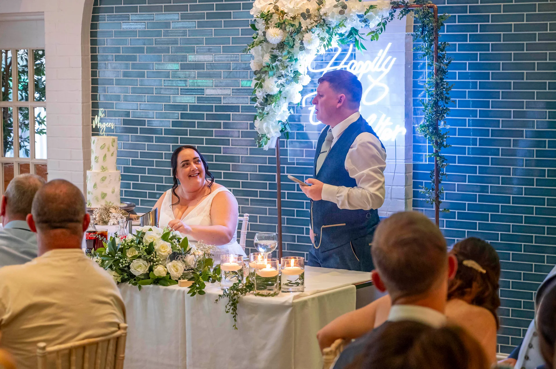 A smiling bride looks up while the groom stands and delivers his wedding speech at the top table in front of a blue tiled wall and illuminated neon sign.