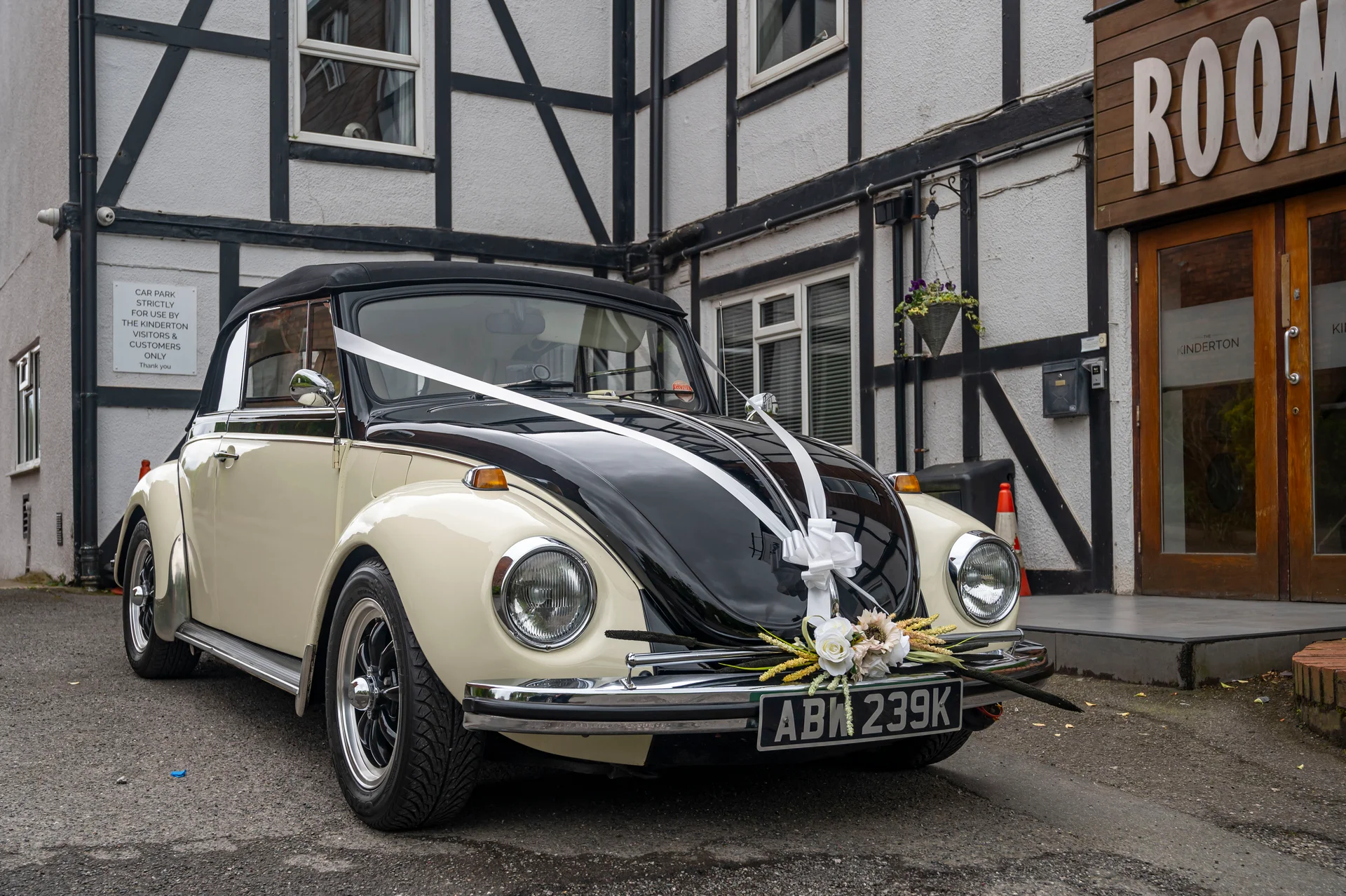 A vintage black and cream Volkswagen Beetle wedding car decorated with white ribbon and floral arrangements parked outside a timber-framed building.