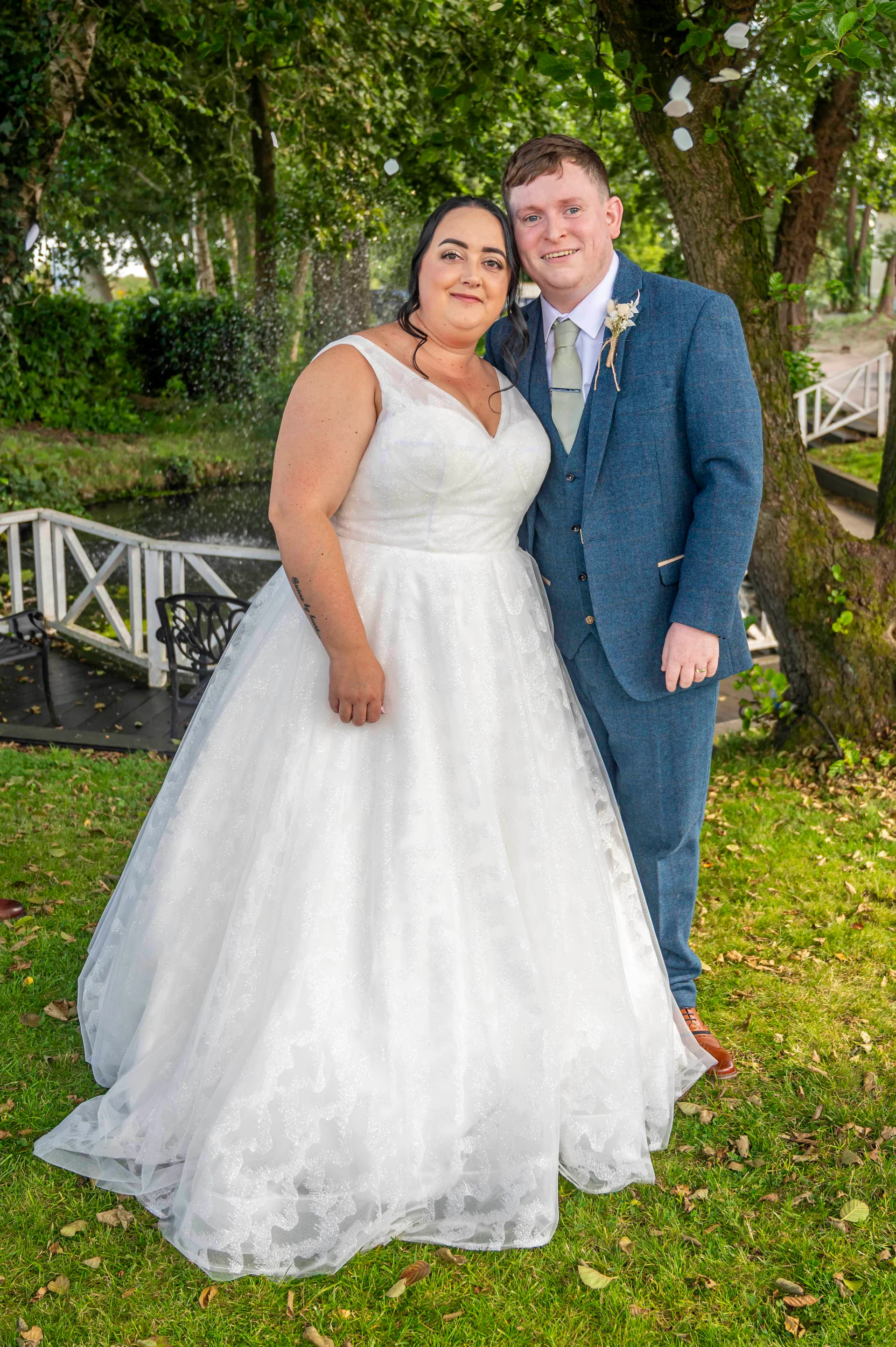 A newlywed couple poses together outdoors on a lush green lawn by a lake, with the bride wearing a sparkling A-line wedding dress and the groom in a stylish blue tweed suit.