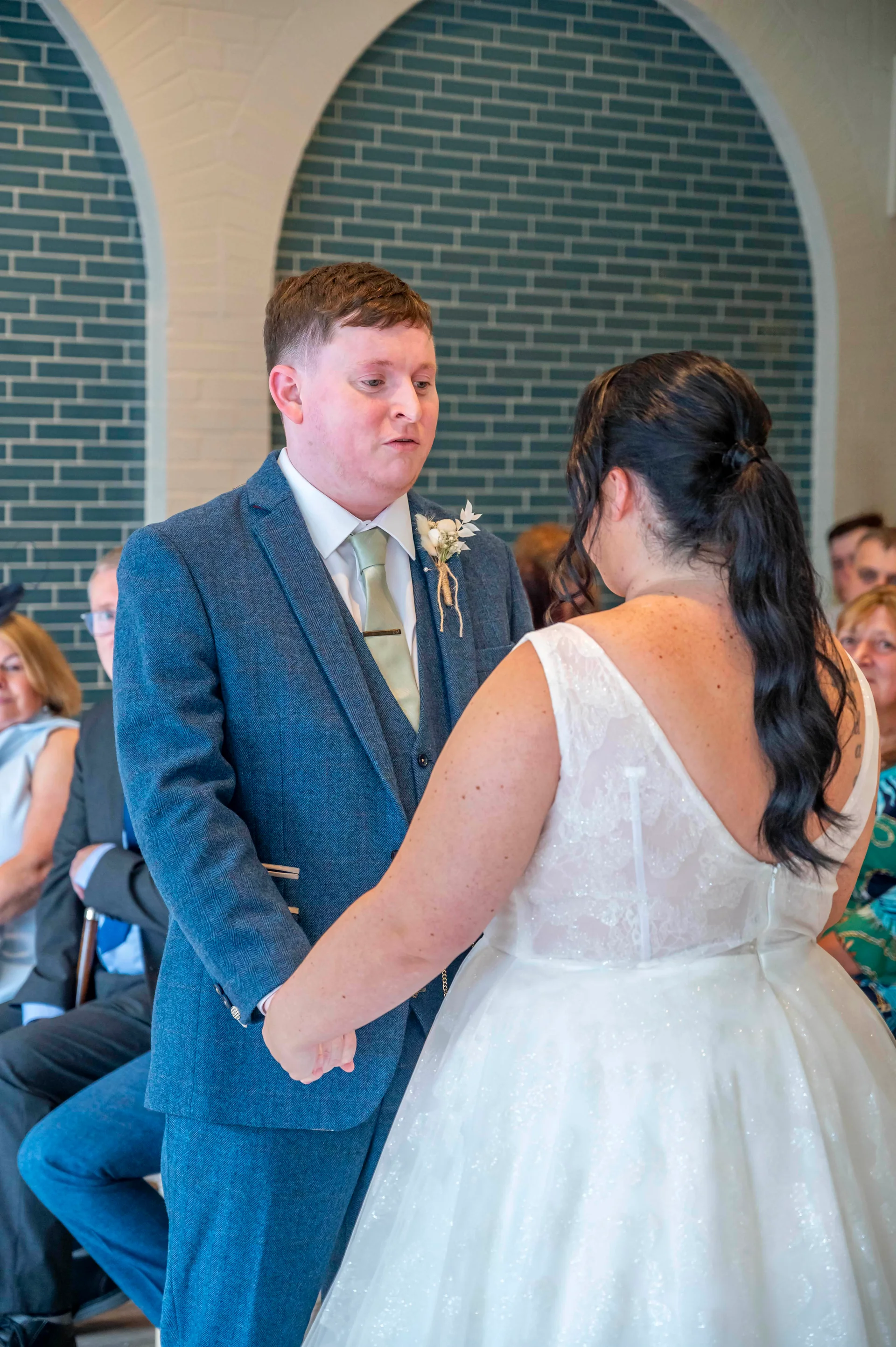 A groom in a blue tweed suit holds hands with his bride wearing a sparkly A-line dress during their indoor civil ceremony.