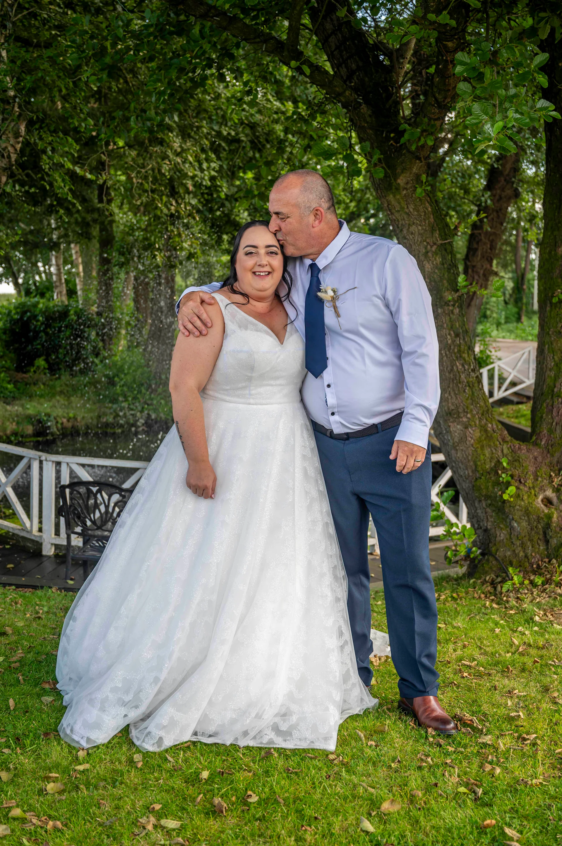 The groom gently kisses the bride on the forehead as they stand together in a lush green garden setting outdoors.