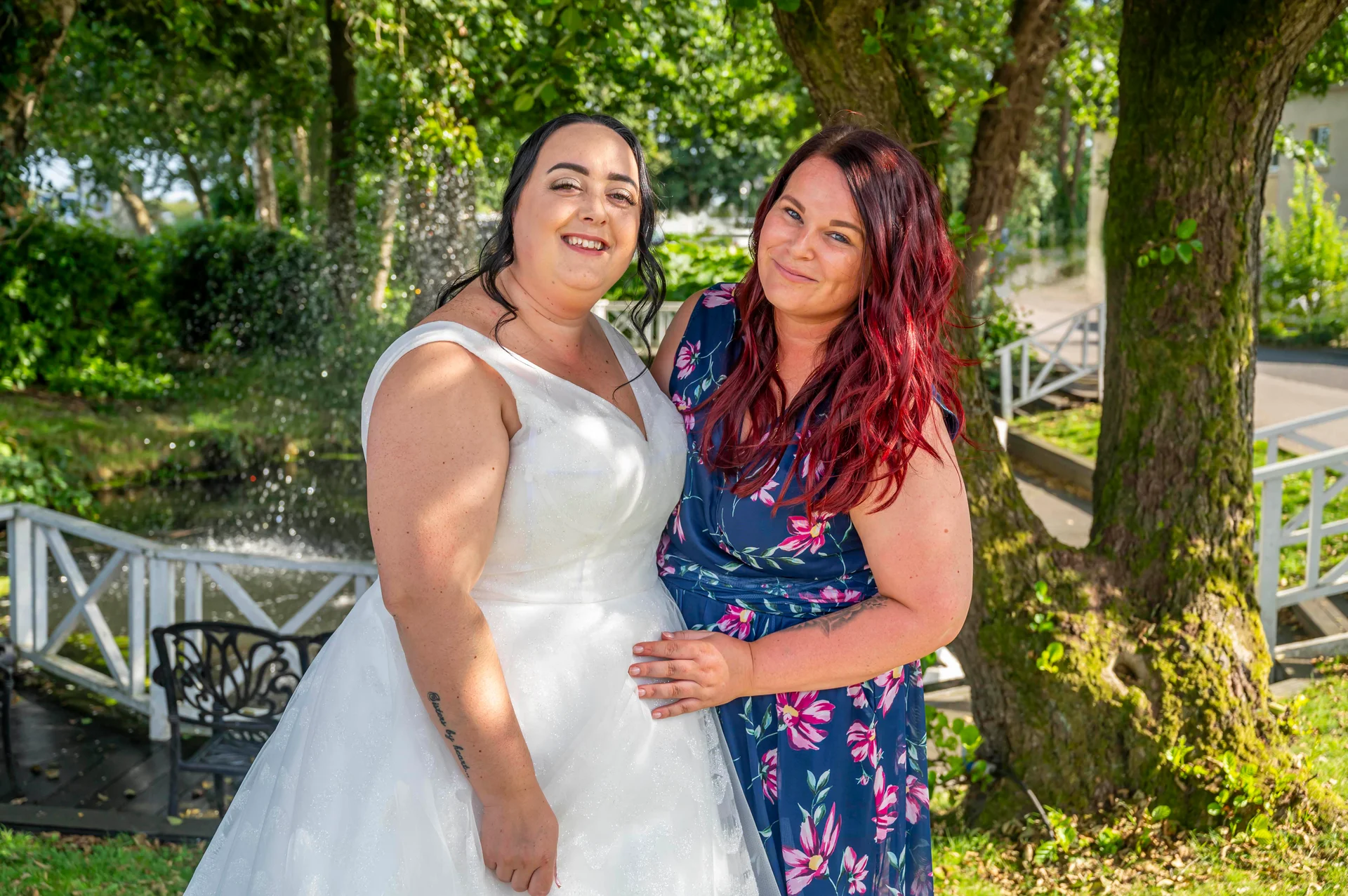 A smiling bride in a white a-line wedding dress stands outdoors next to a guest in a blue floral dress near a fountain and trees.