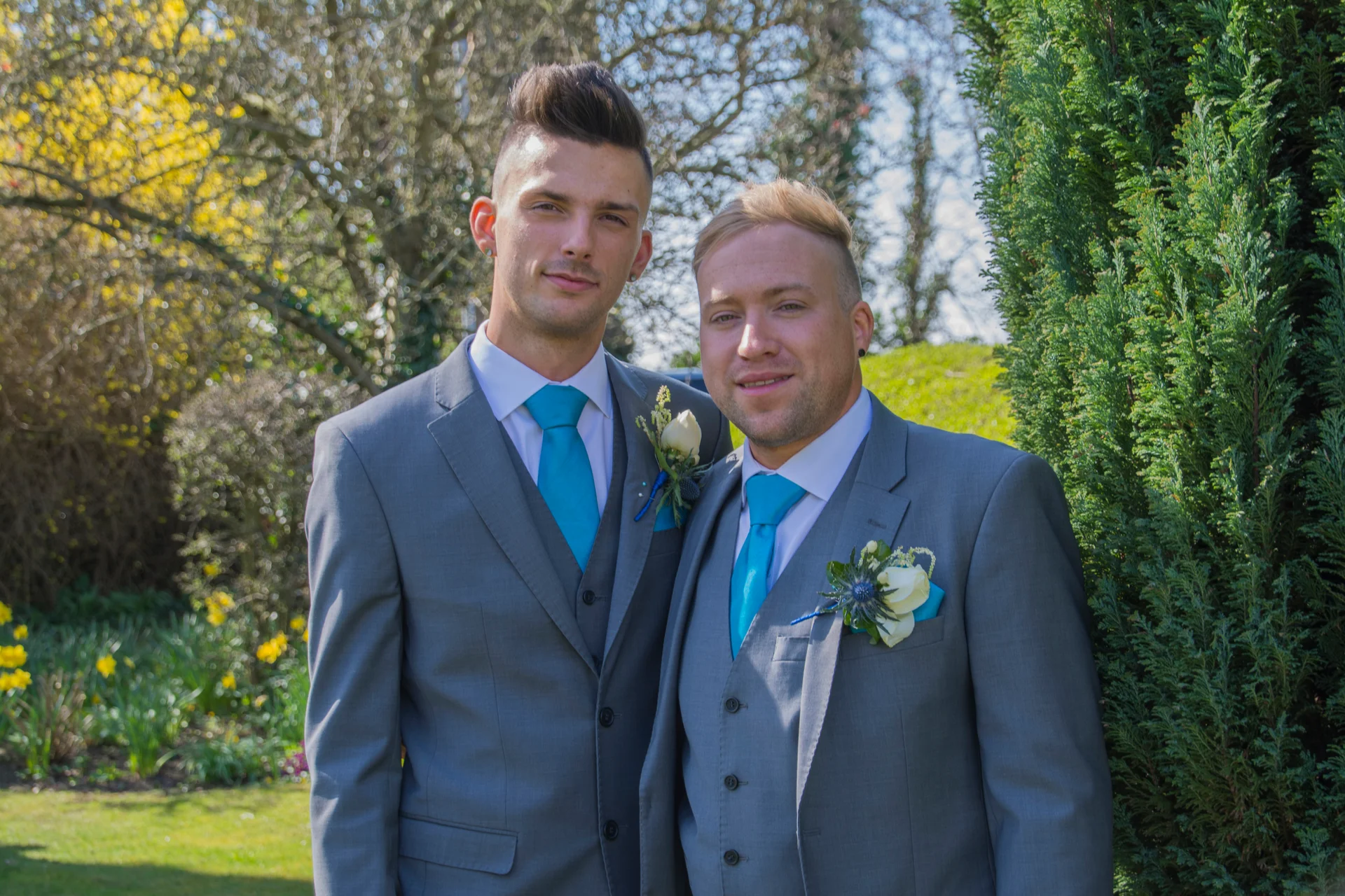 Two grooms stand side by side in matching grey three-piece suits with bright blue ties and white floral buttonholes in an outdoor garden setting.