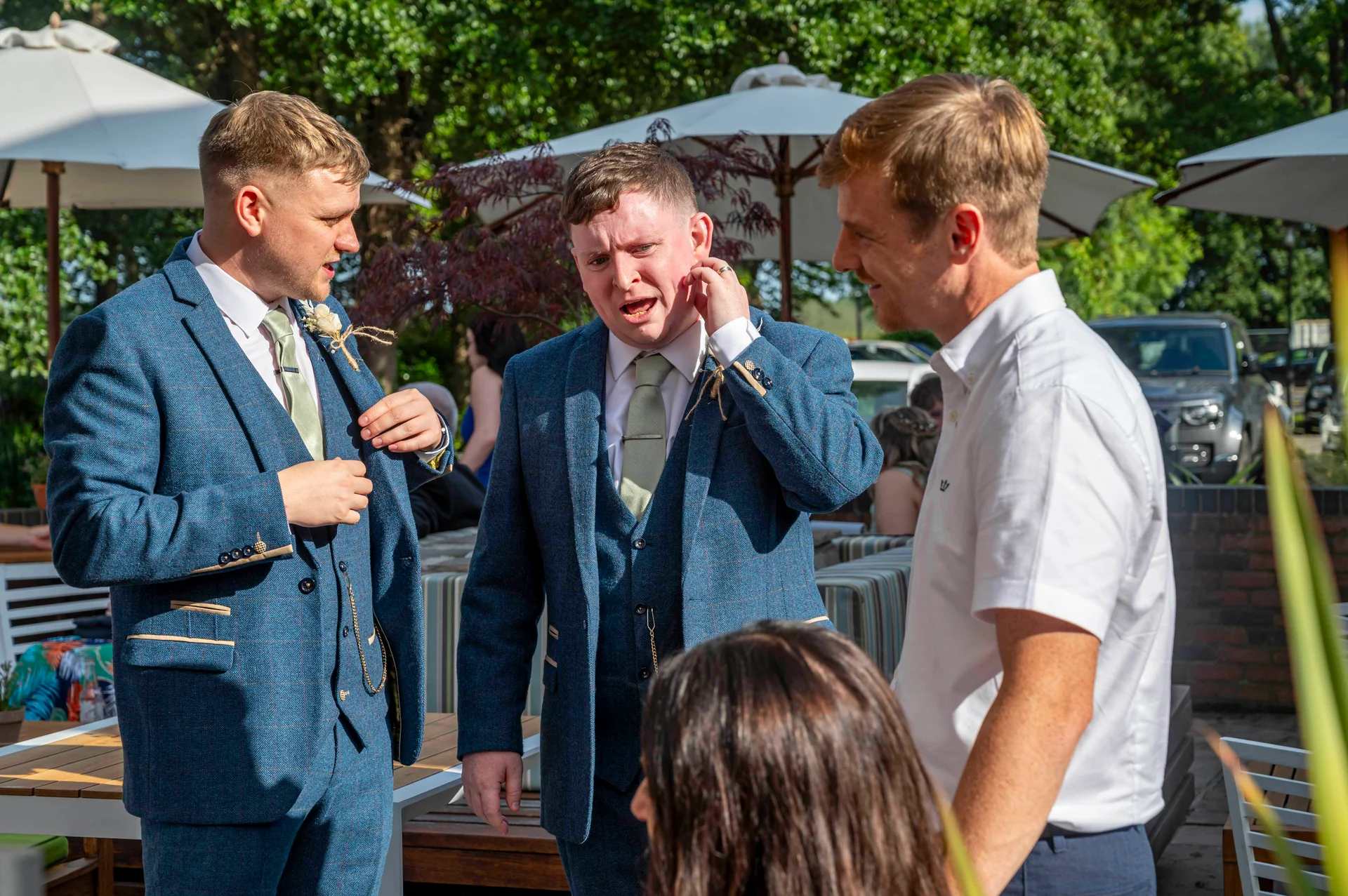 Groomsmen in blue tweed suits chatting and laughing together outdoors during a sunny wedding reception.