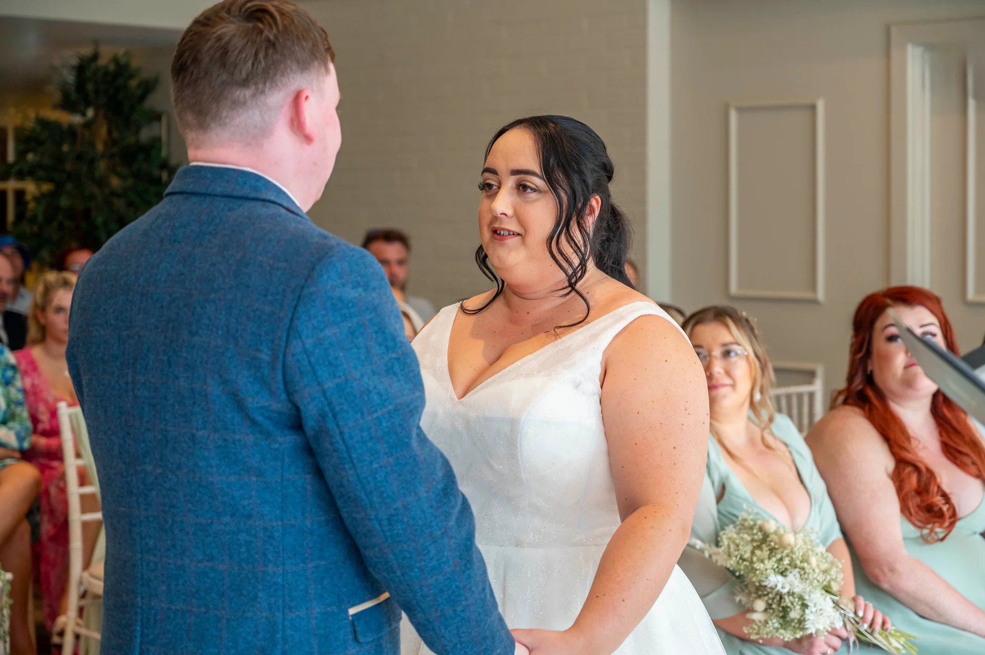 The bride and groom exchange vows during an indoor civil ceremony, with the groom wearing a blue checked suit and the bride in a sparkly v-neck gown.