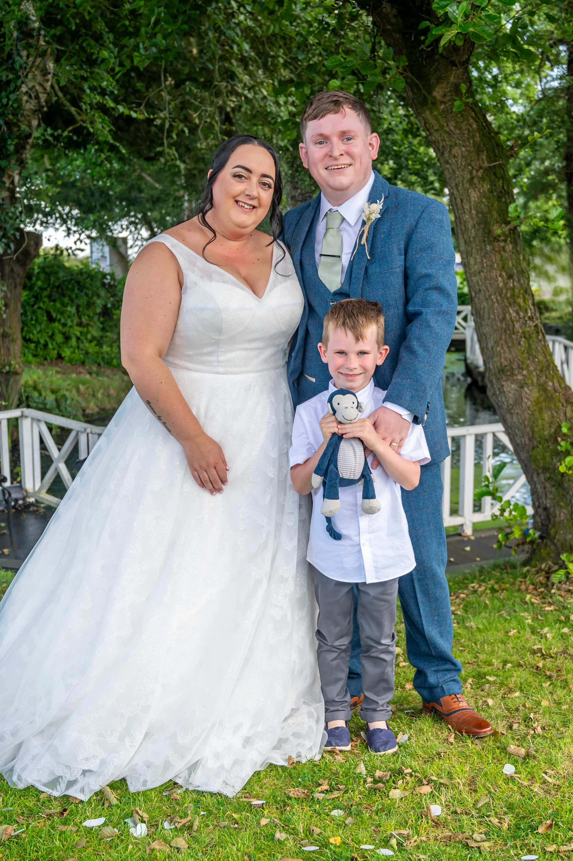 A smiling bride in a white A-line gown and groom in a blue tweed suit pose outdoors with a young page boy holding a small toy.