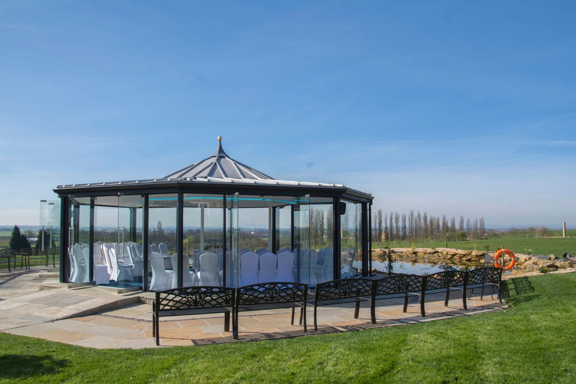A modern glasshouse wedding ceremony pavilion stands outdoors beside a tranquil lake under a clear blue sky.
