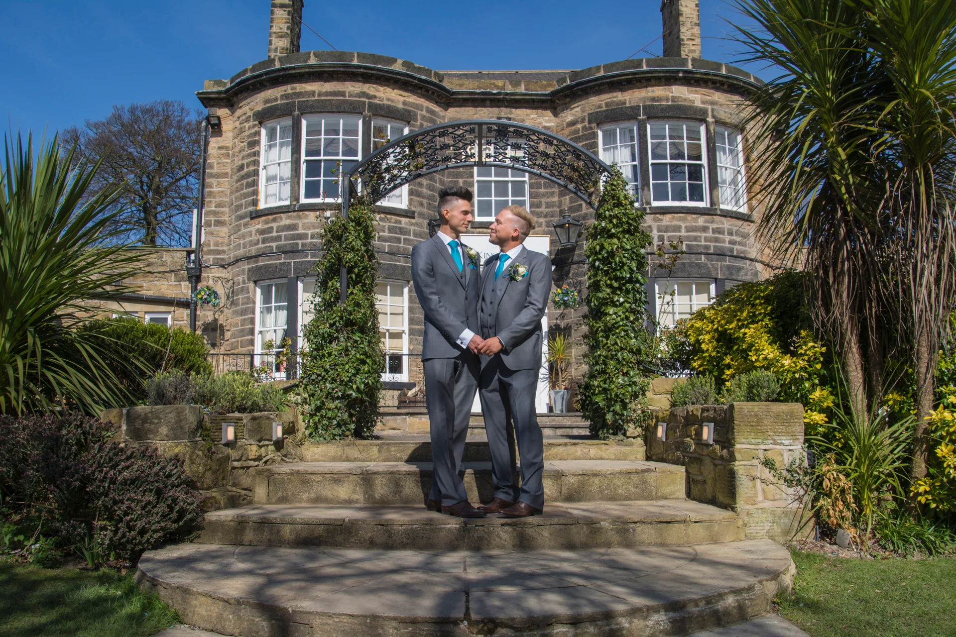 Two grooms in matching grey suits stand hand in hand under a stone archway outside a historic country house on a sunny day.