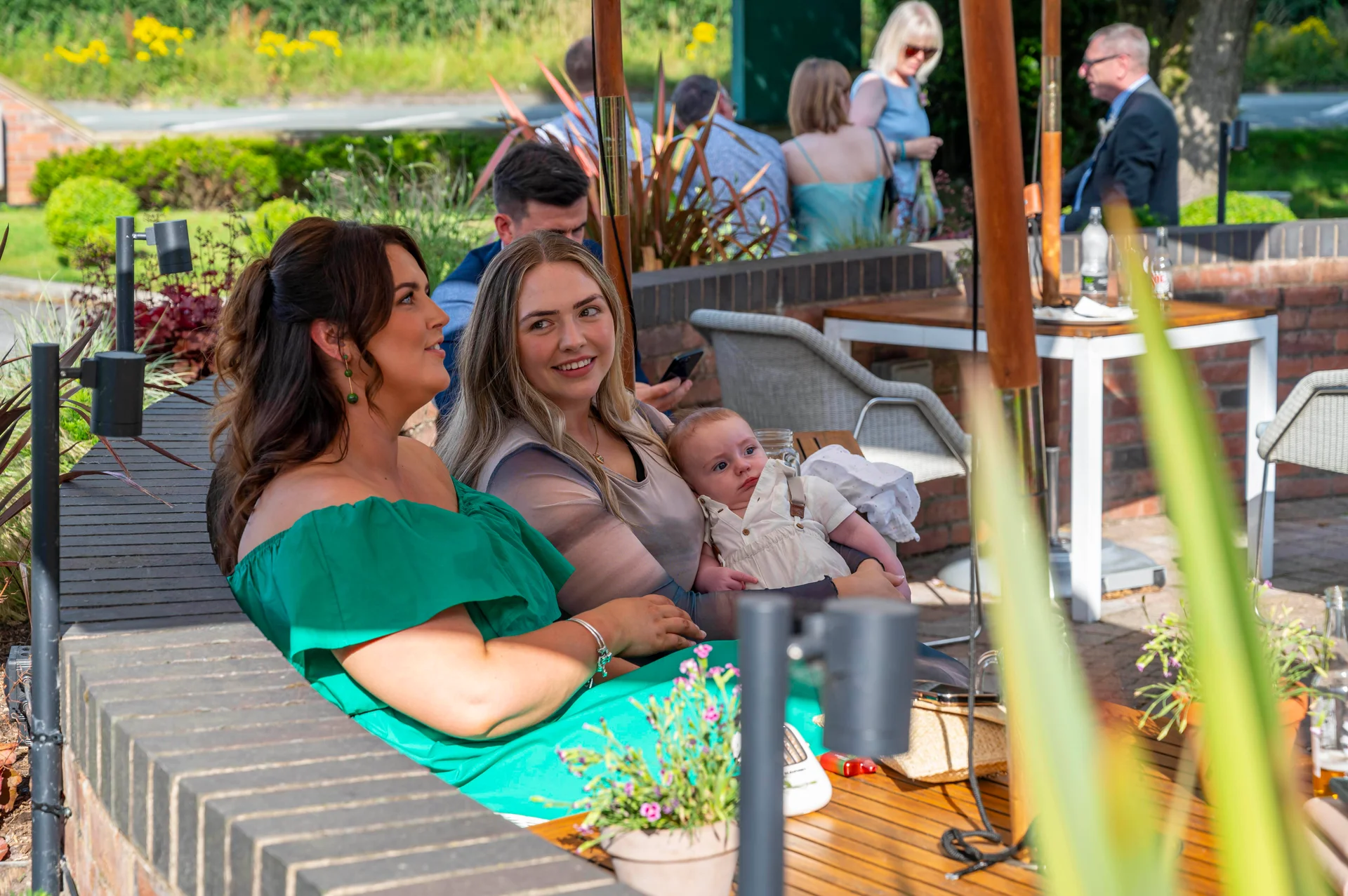 Two women relax together on an outdoor terrace during a wedding reception, with one holding a baby while other guests mingle in the background under bright sunshine.