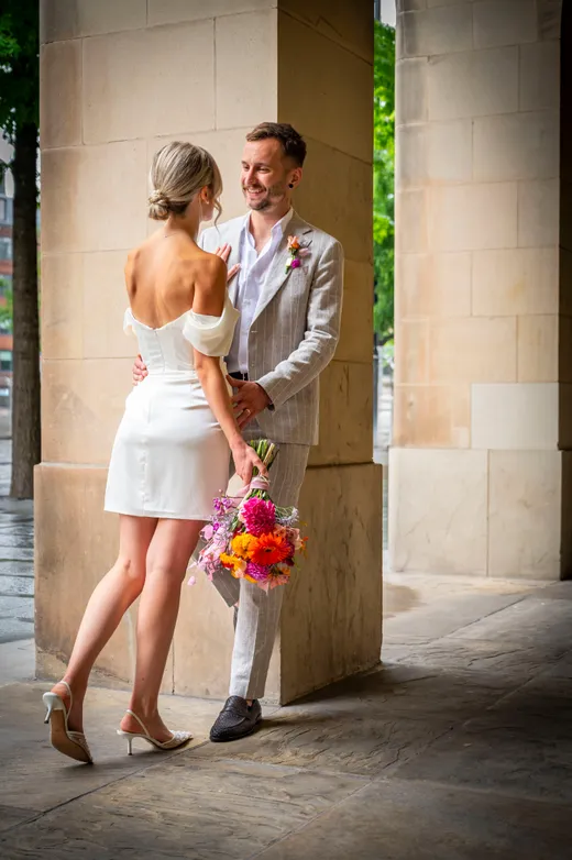 A stylish newlywed couple embrace near stone pillars, with the bride wearing a short white satin dress and holding a vibrant colourful bouquet while the groom wears a light grey patterned suit.
