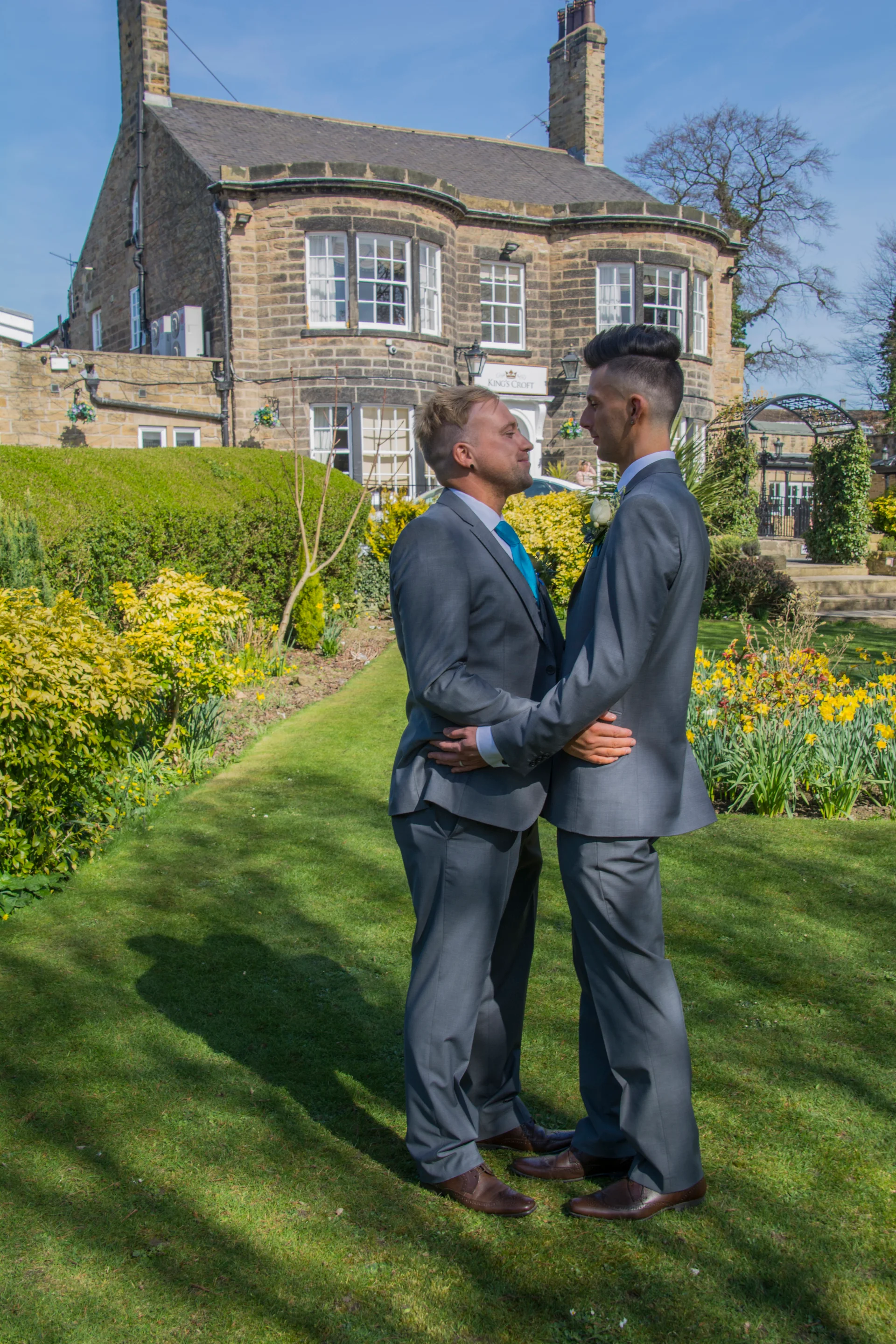 Two grooms in matching grey suits embrace in the manicured gardens of a historic country house on a bright spring day.