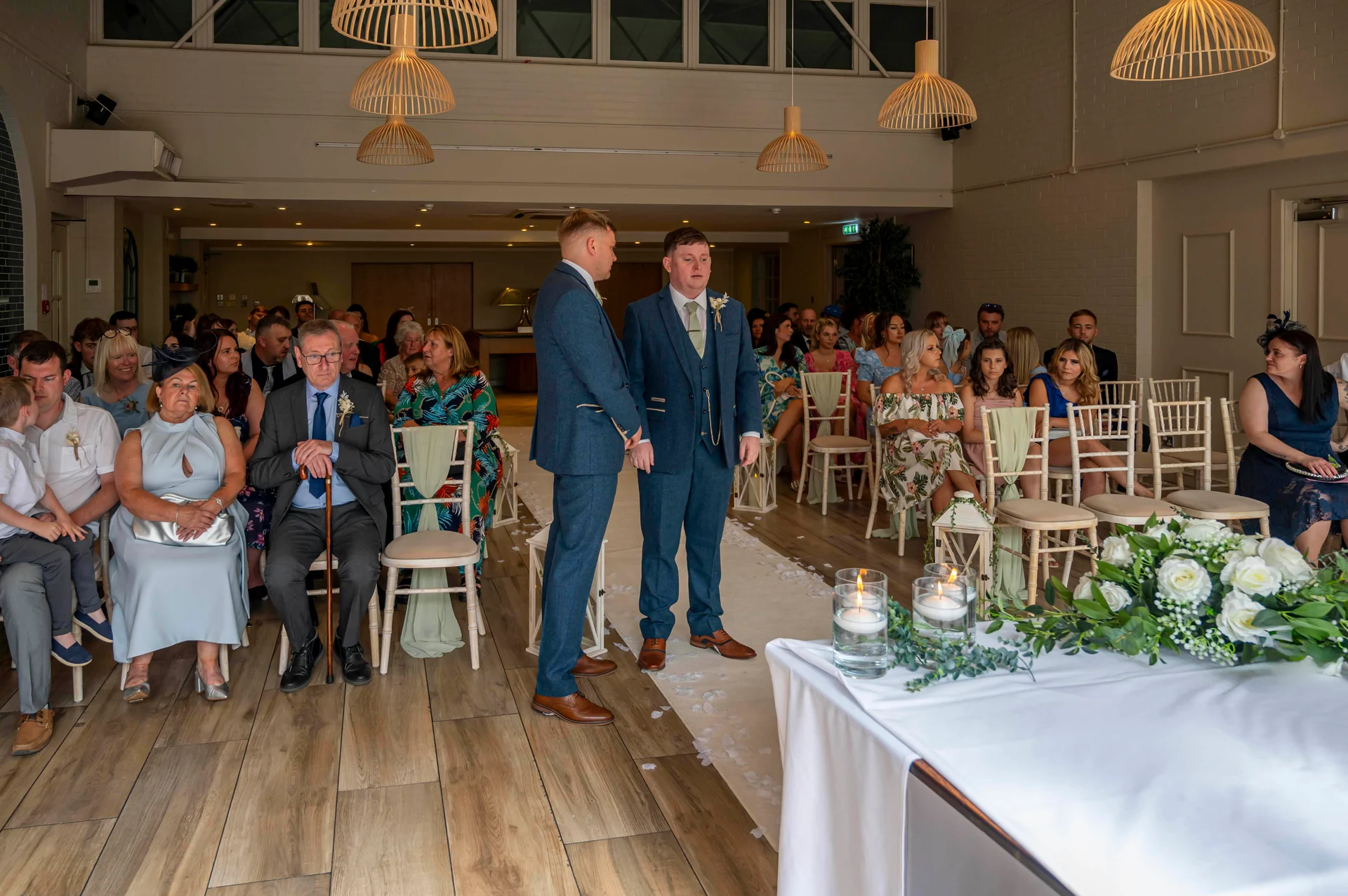 Two grooms in matching blue suits stand at the front of a light-filled ceremony room, surrounded by seated wedding guests and floral arrangements.