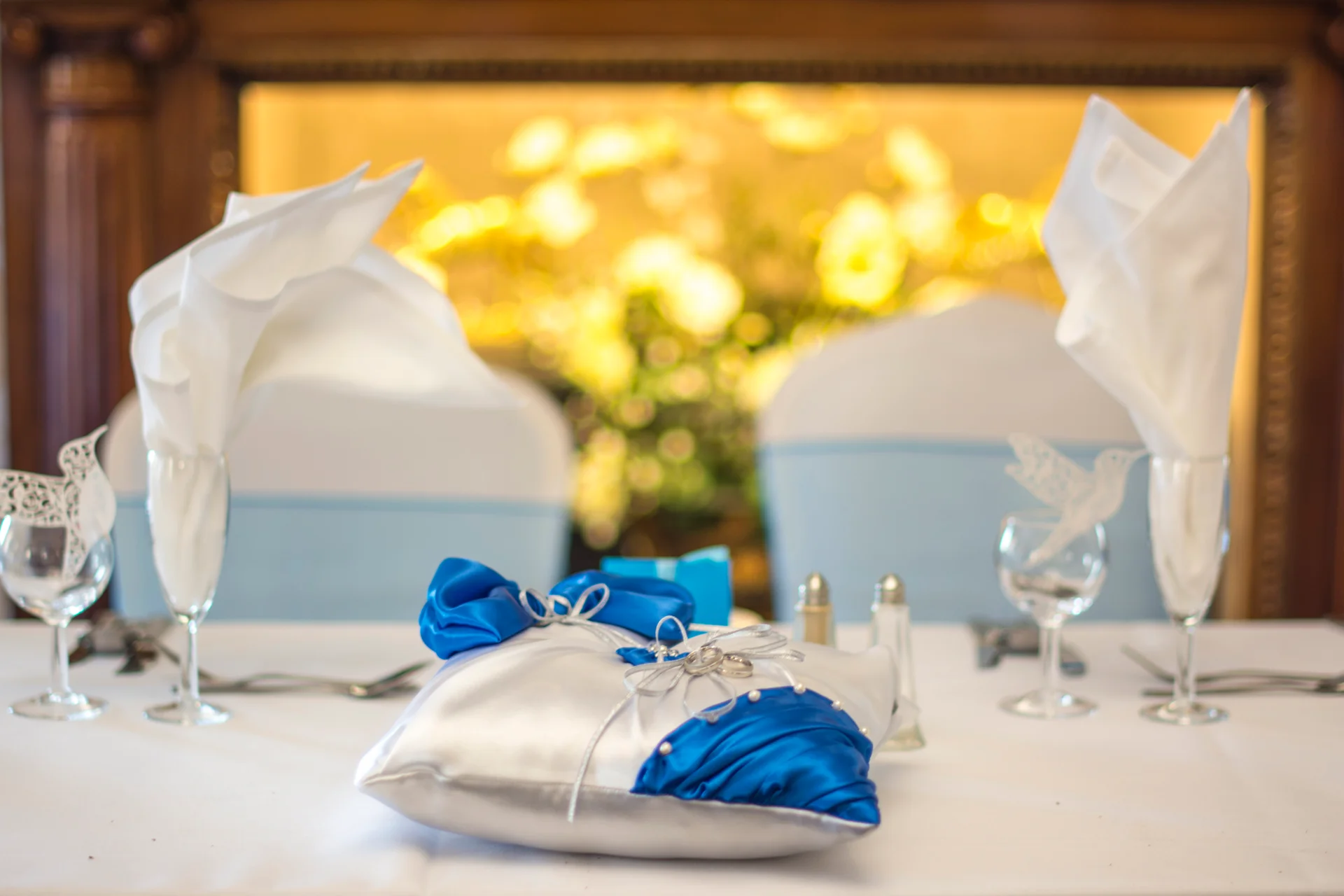 A decorative silver and blue satin ring pillow rests on a neatly set wedding reception table featuring folded white napkins and crystal glassware.