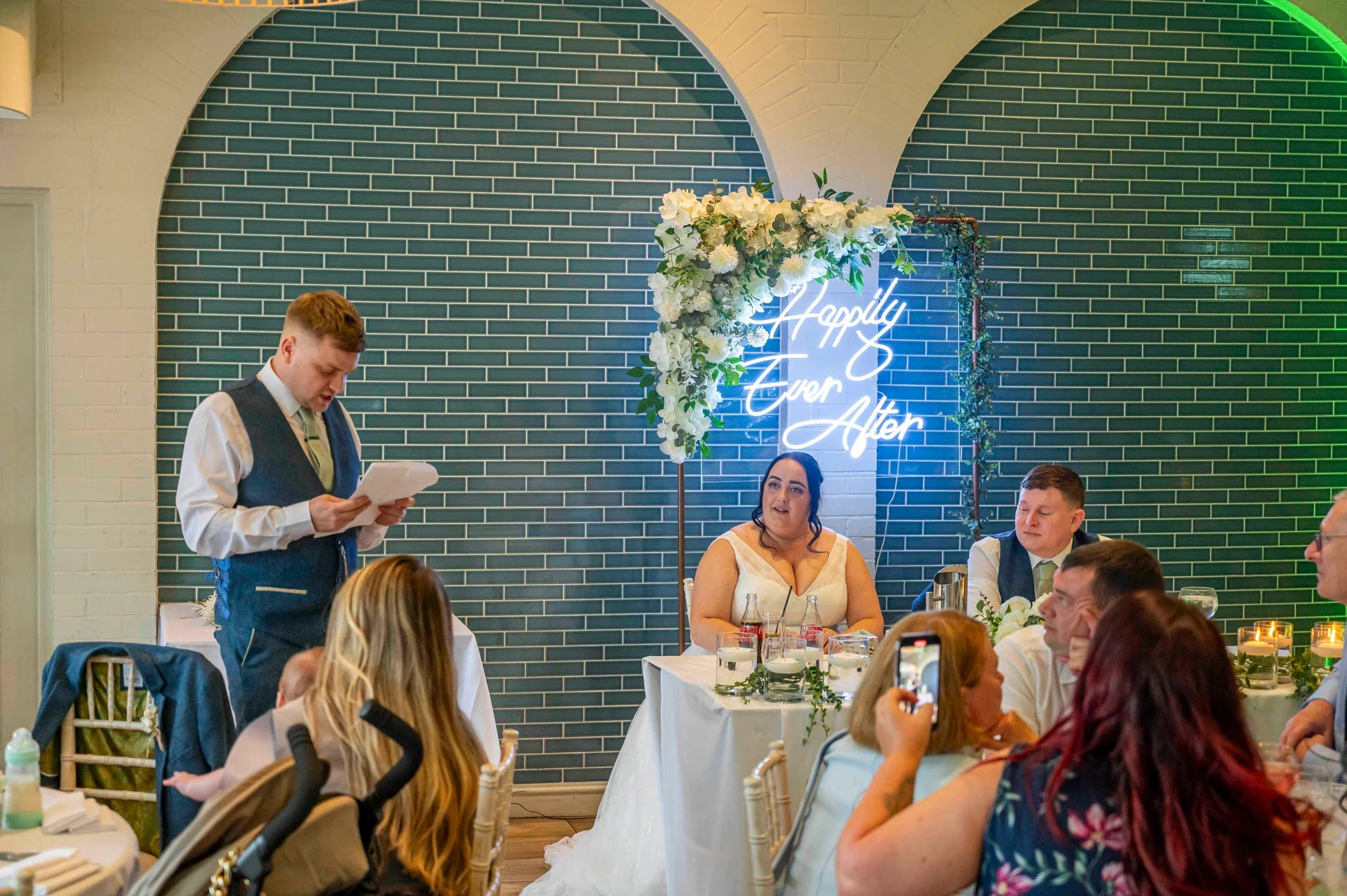 A groom delivers a speech holding notes at the top table, while the bride and wedding guests listen in front of a striking teal tiled wall featuring a neon sign and floral arch.
