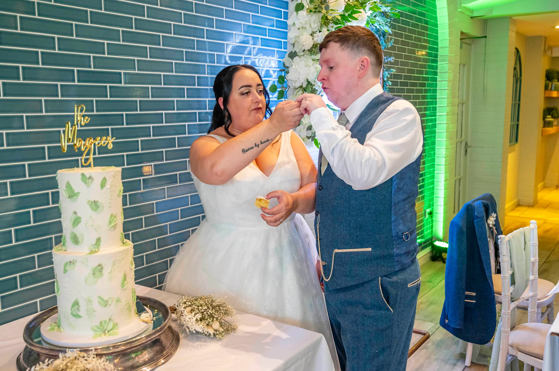 A bride and groom feed each other a bite of their two-tier green-and-white wedding cake in front of a turquoise tiled wall.