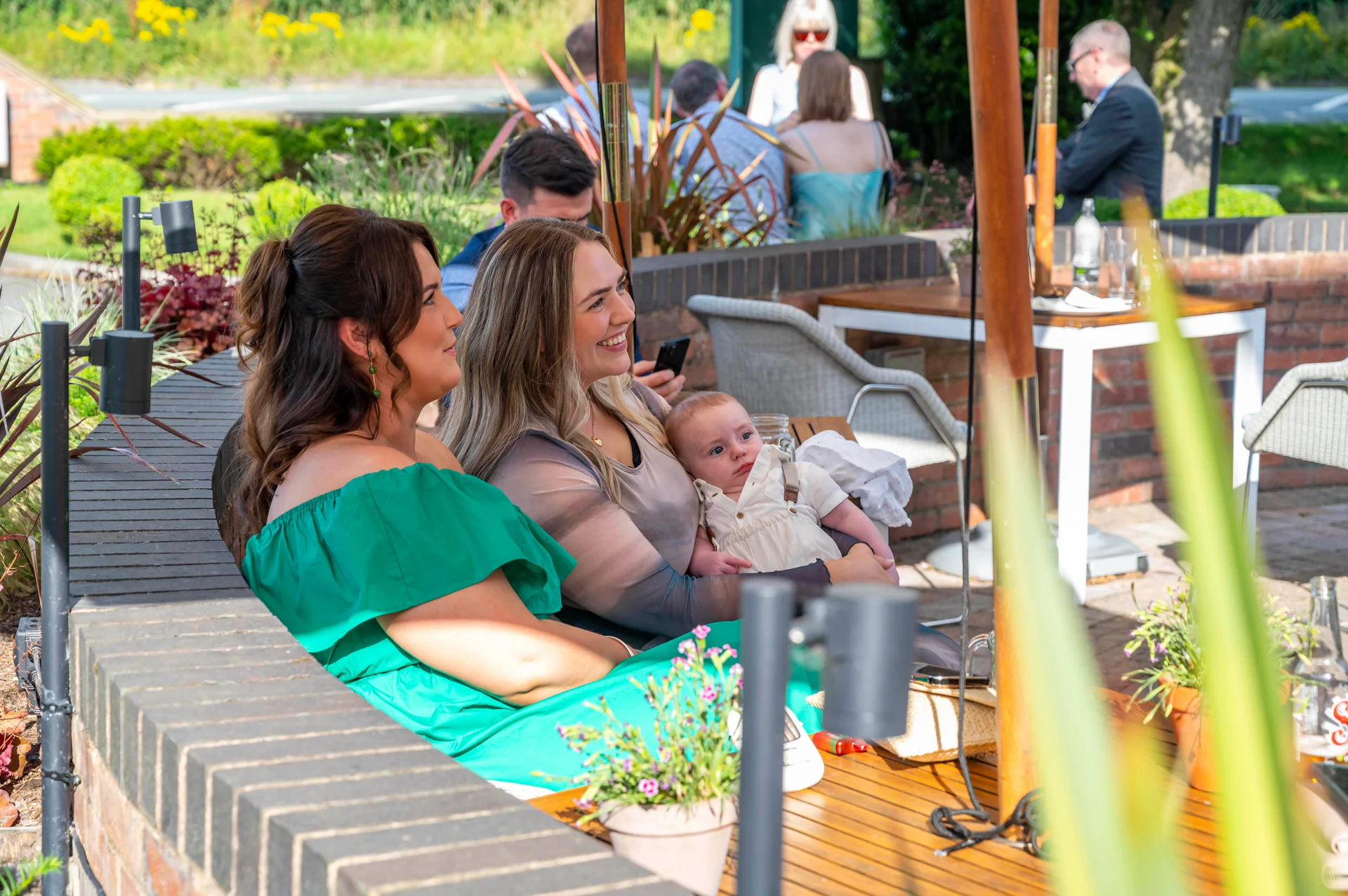 Wedding guests relaxing outdoors at an outdoor reception, with a baby in a white outfit held by a woman in a green dress.