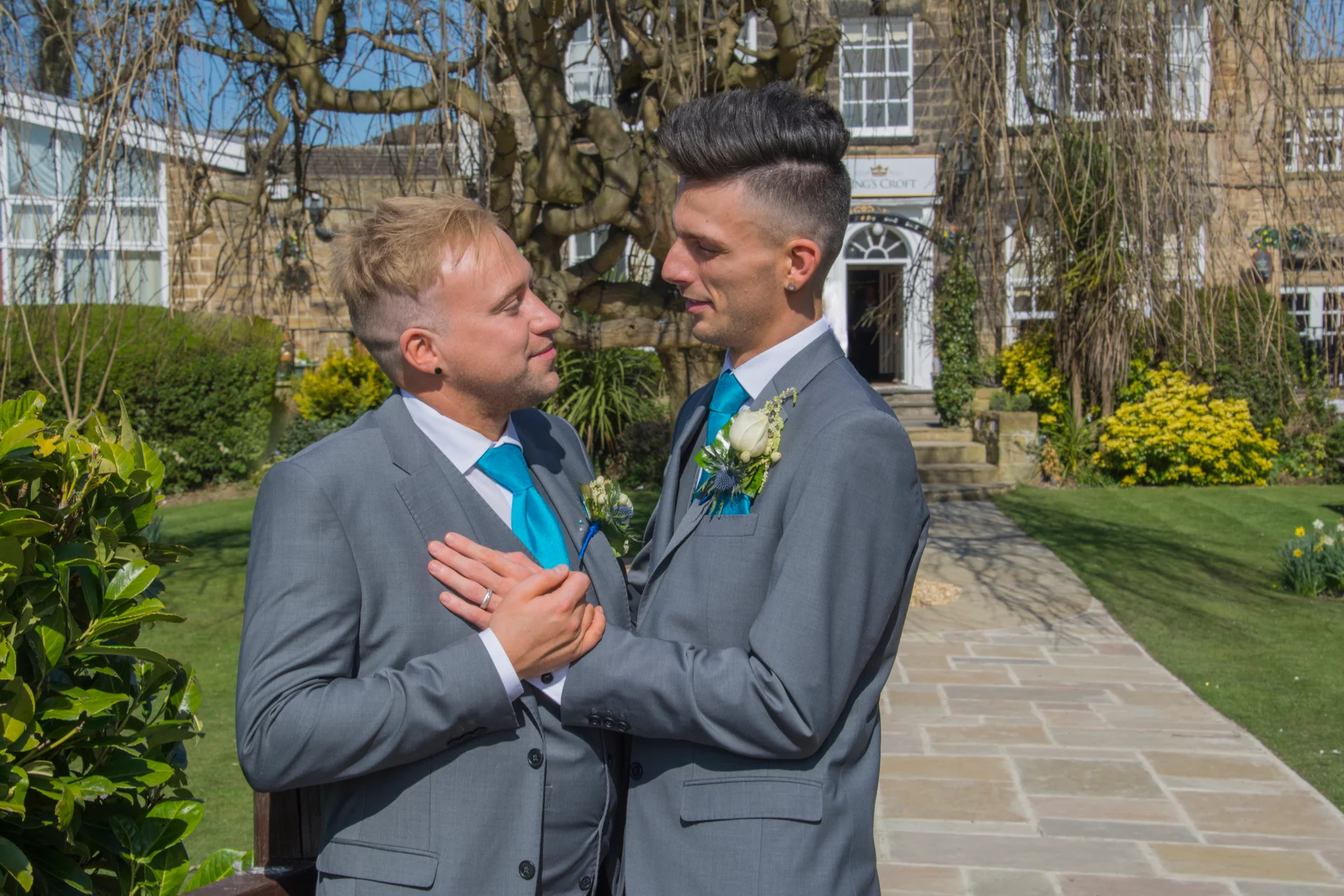 Two grooms in matching grey suits and bright blue ties affectionately embrace outdoors in the manicured gardens of a stately country house venue.