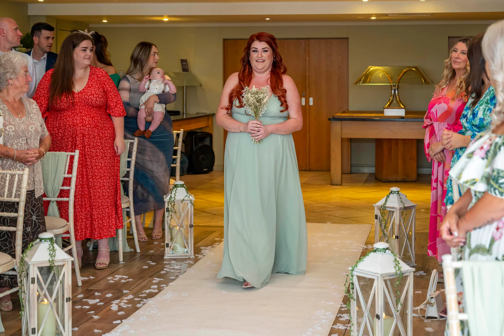 A smiling bride with vibrant red hair walks down the aisle in a sage green gown, holding a bouquet, as guests watch from seating adorned with lanterns and scattered petals.