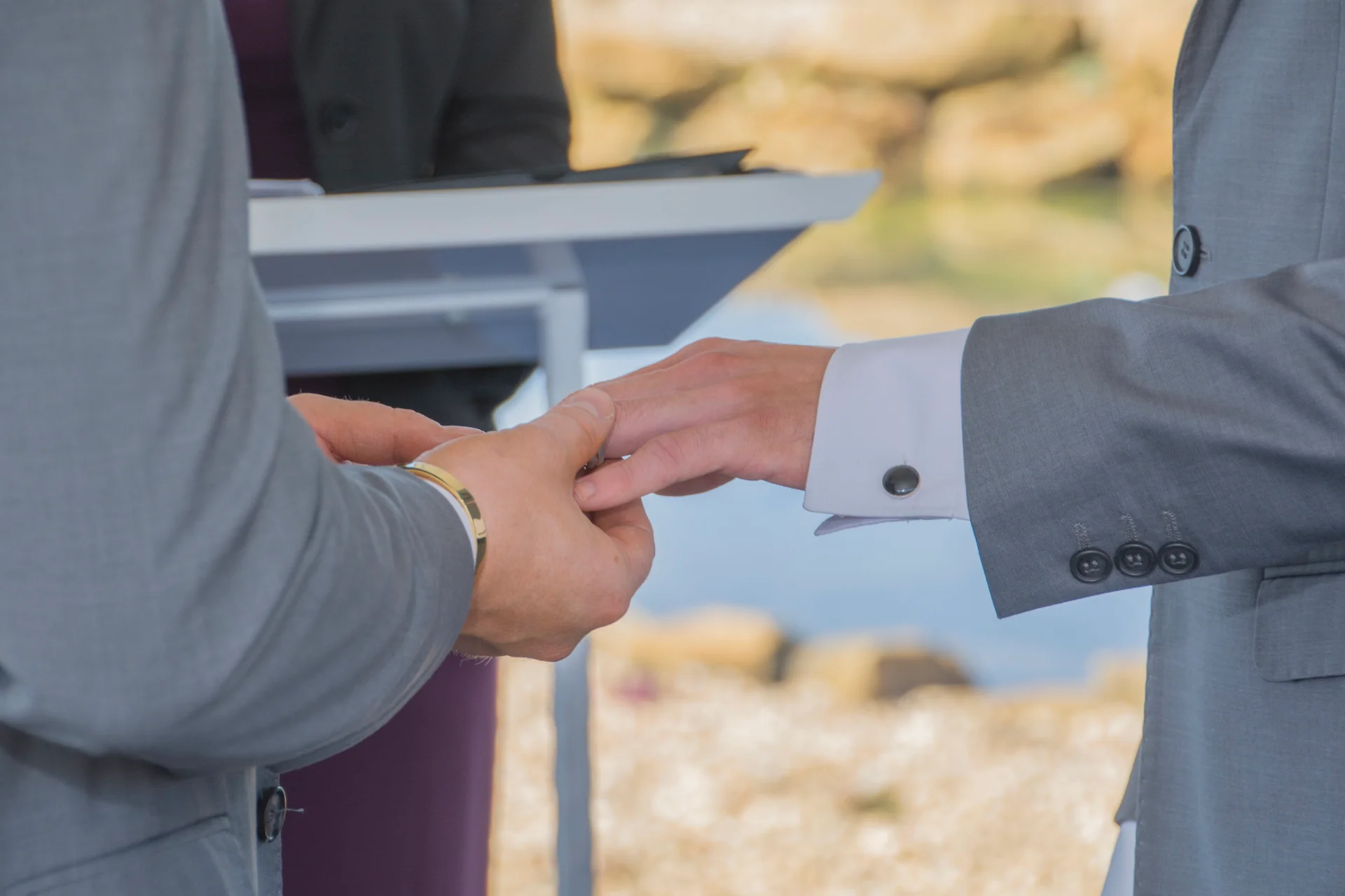 Two grooms in matching grey suits exchange wedding bands during an outdoor civil ceremony.