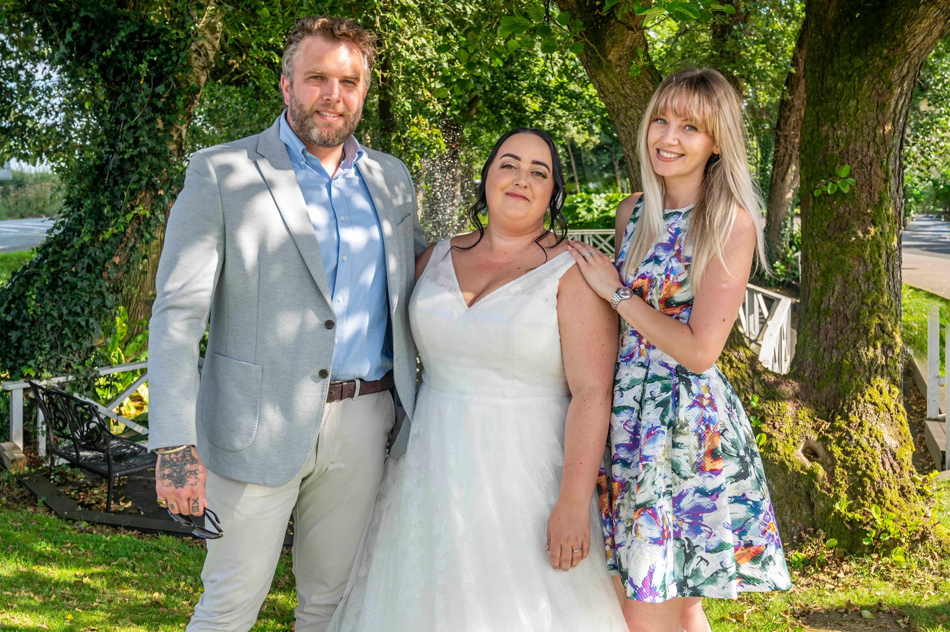 A smiling bride in a white tulle gown stands outdoors between a man in a grey suit and a bridesmaid in a floral dress during a summer wedding.