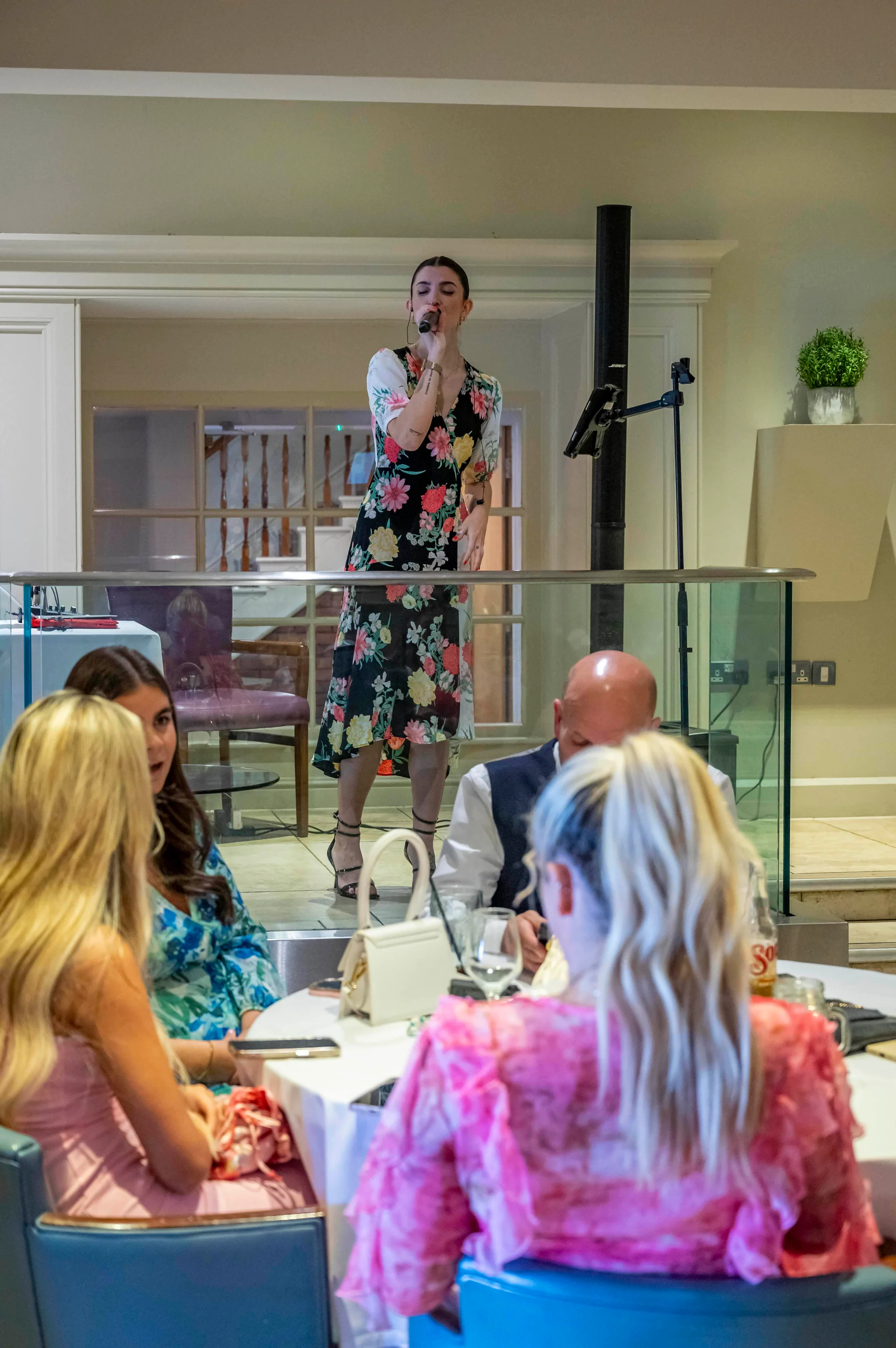 A female singer performs into a microphone on a raised platform during an indoor wedding reception while guests sit at nearby tables.