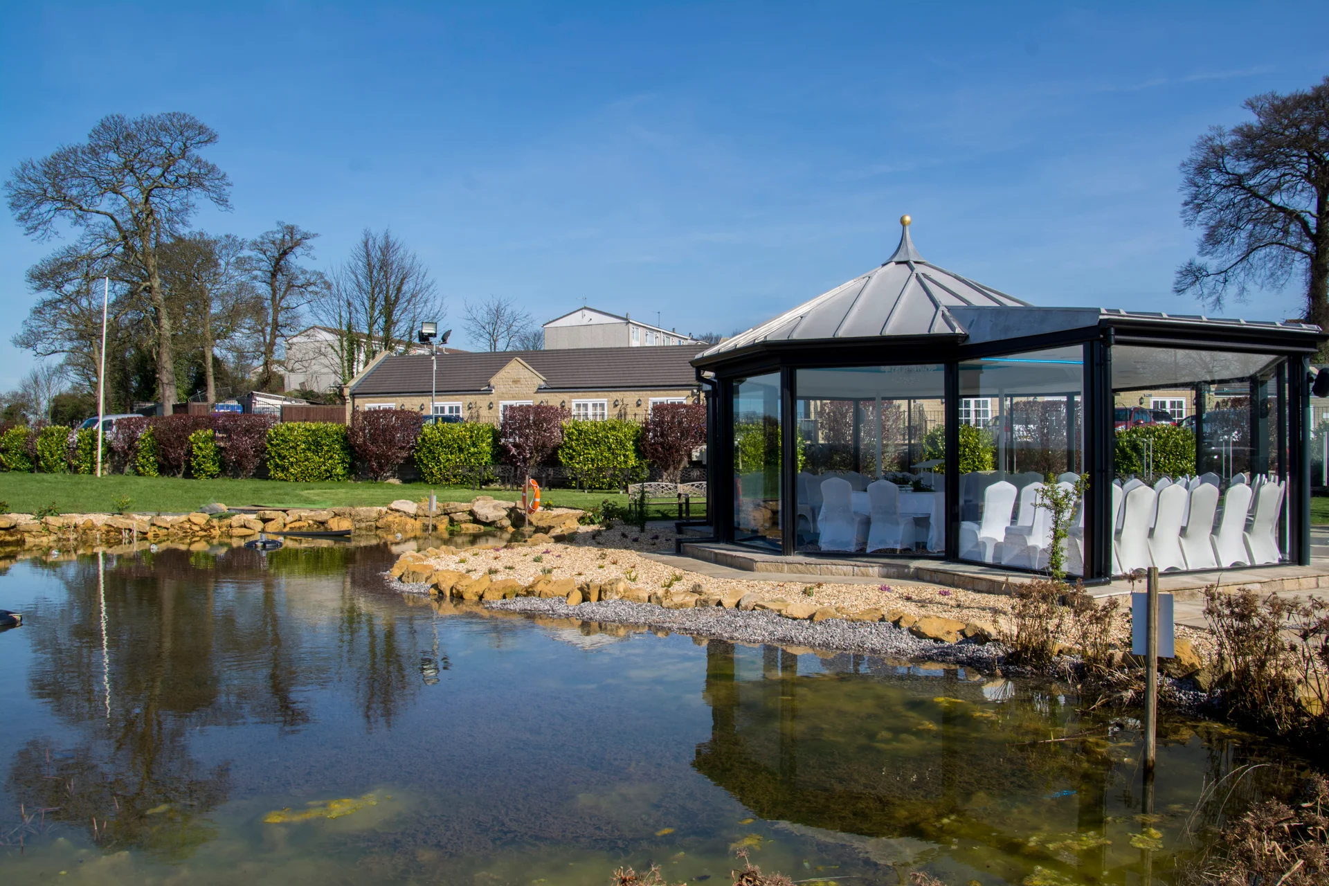 A modern glasshouse wedding pavilion sits beside a tranquil lake under a clear blue sky on a sunny day.