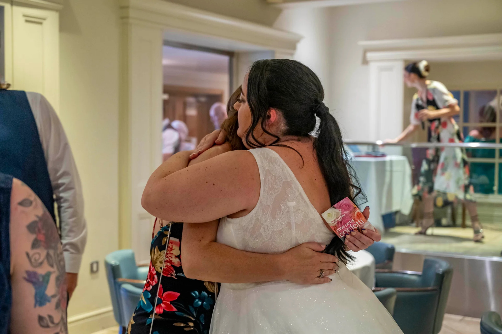 A bride in a white lace dress with a low ponytail is captured in a warm, emotional embrace with a guest inside an indoor venue.