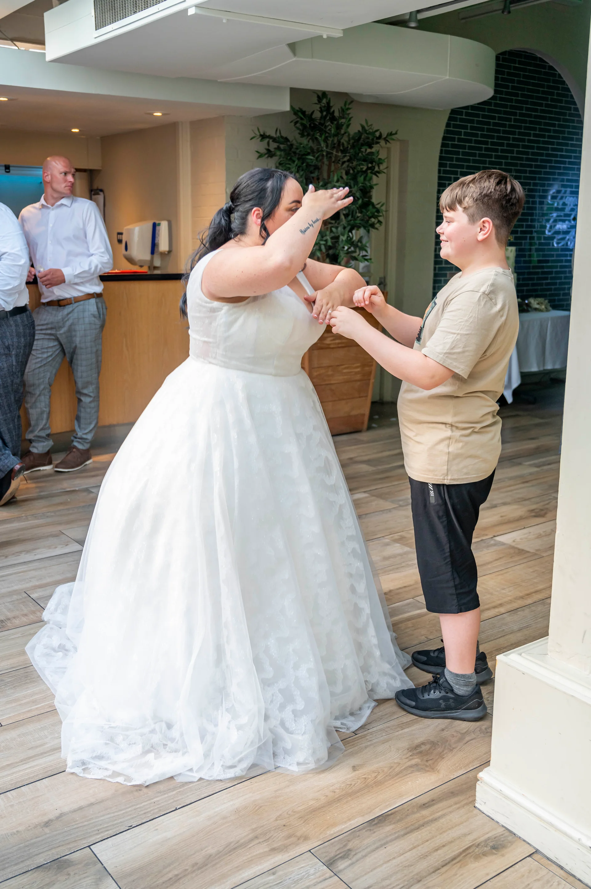 A bride in a white patterned a line wedding dress shares a lighthearted moment with a young guest indoors.