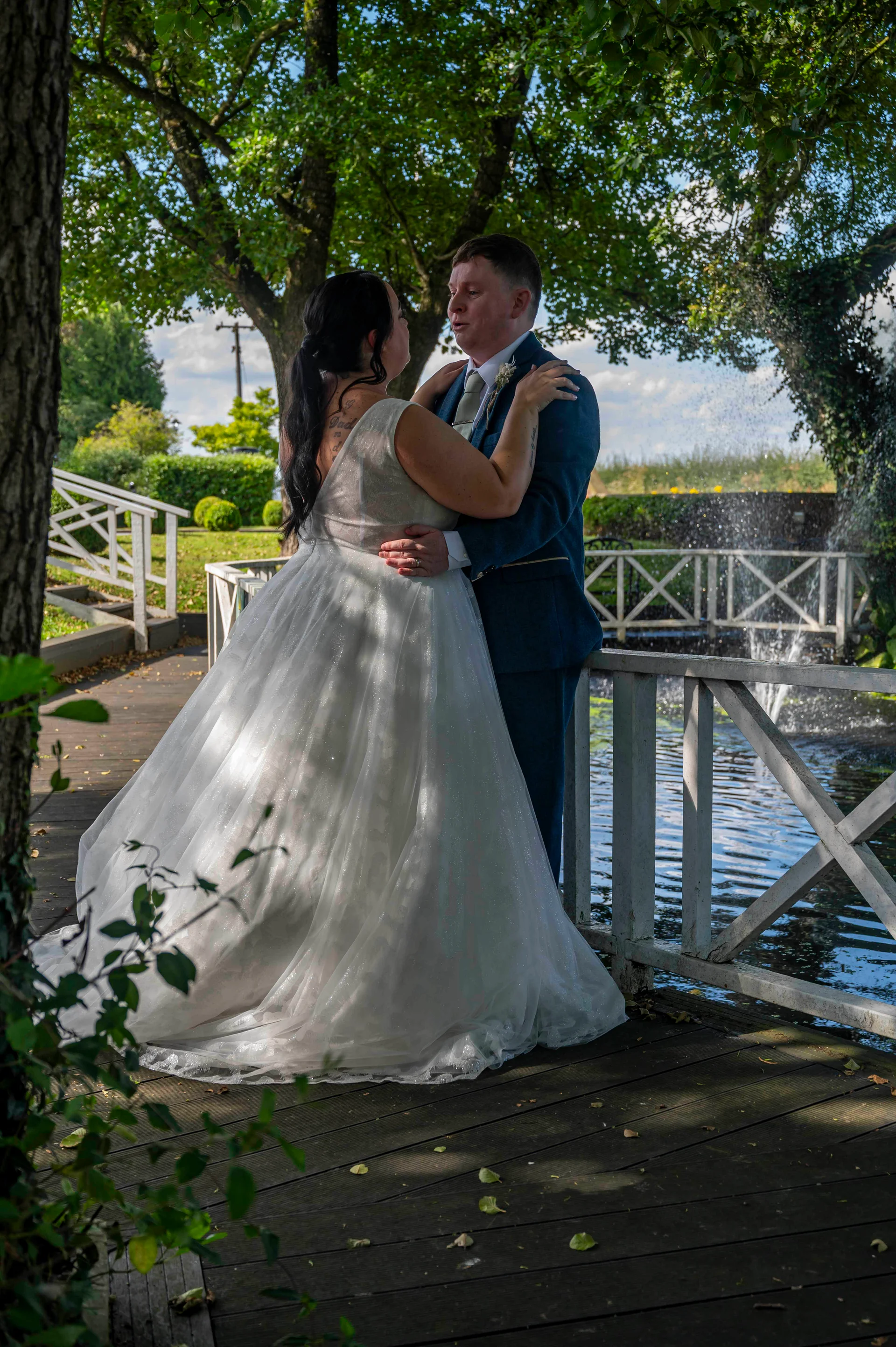 A newlywed couple embraces on a wooden bridge near a sparkling fountain during their outdoor wedding portrait session.